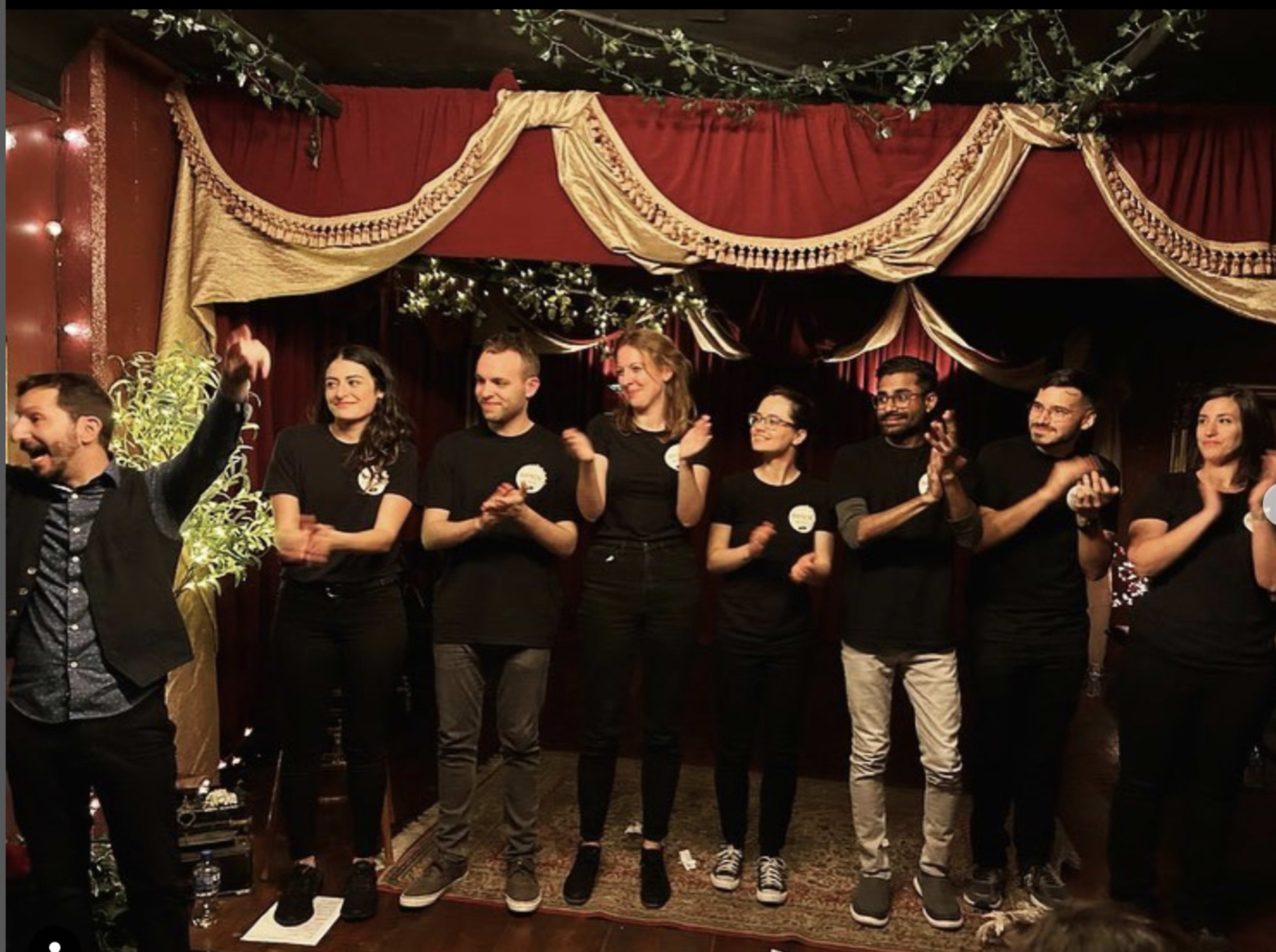 A group of eight performers stands on stage, dressed in black shirts, applauding with a backdrop of festive decorations and dim lighting.