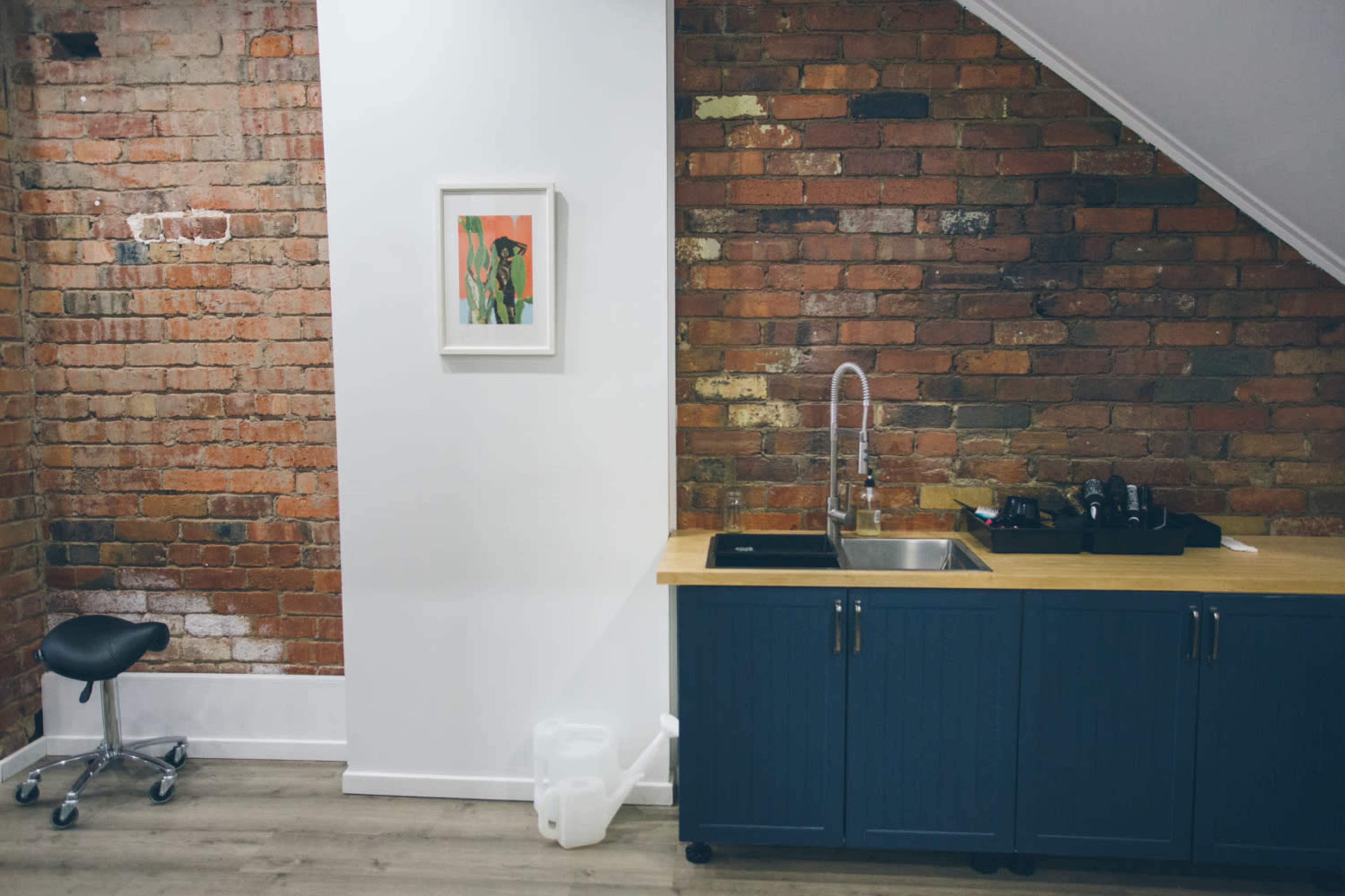A small kitchen area with a brick wall, a stainless steel sink, and a blue cabinet beneath a framed artwork on the wall.
