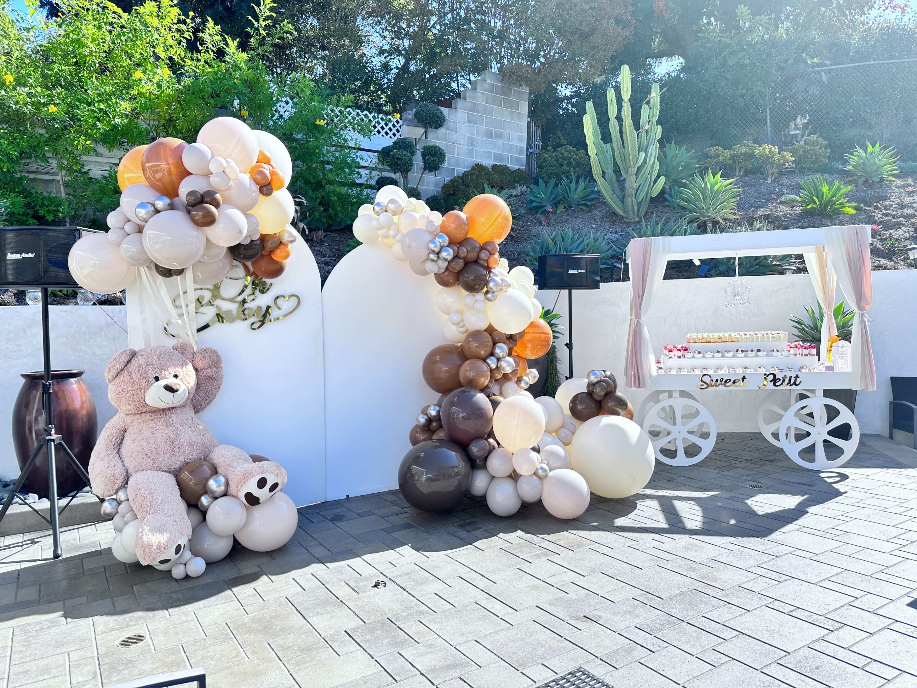 The scene features a decorated outdoor space with a balloon arch on one side and a cake cart on the other, complemented by a large teddy bear.