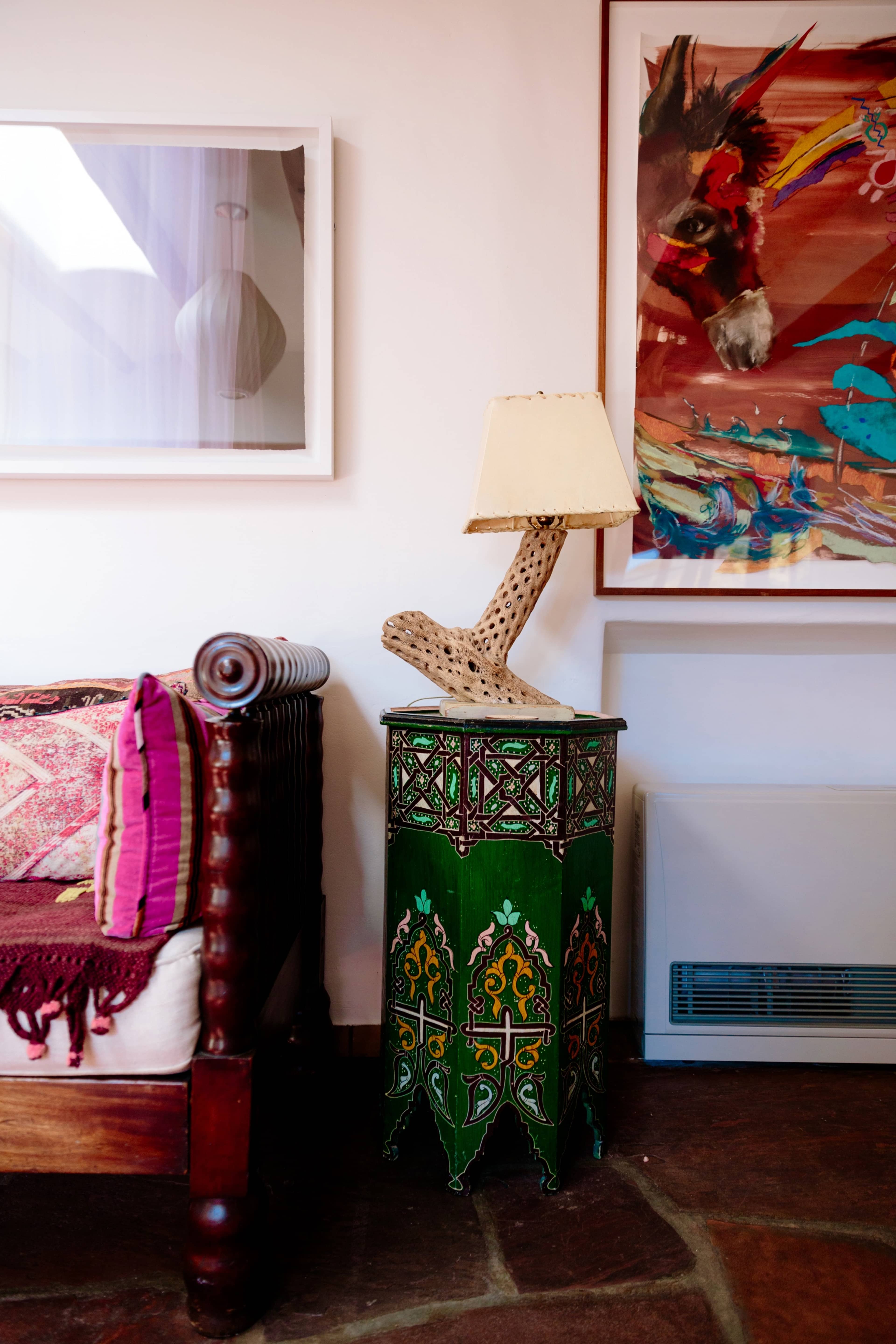 A wooden side table with intricate green designs holds a lamp, beside a patterned sofa and framed artwork on the wall.