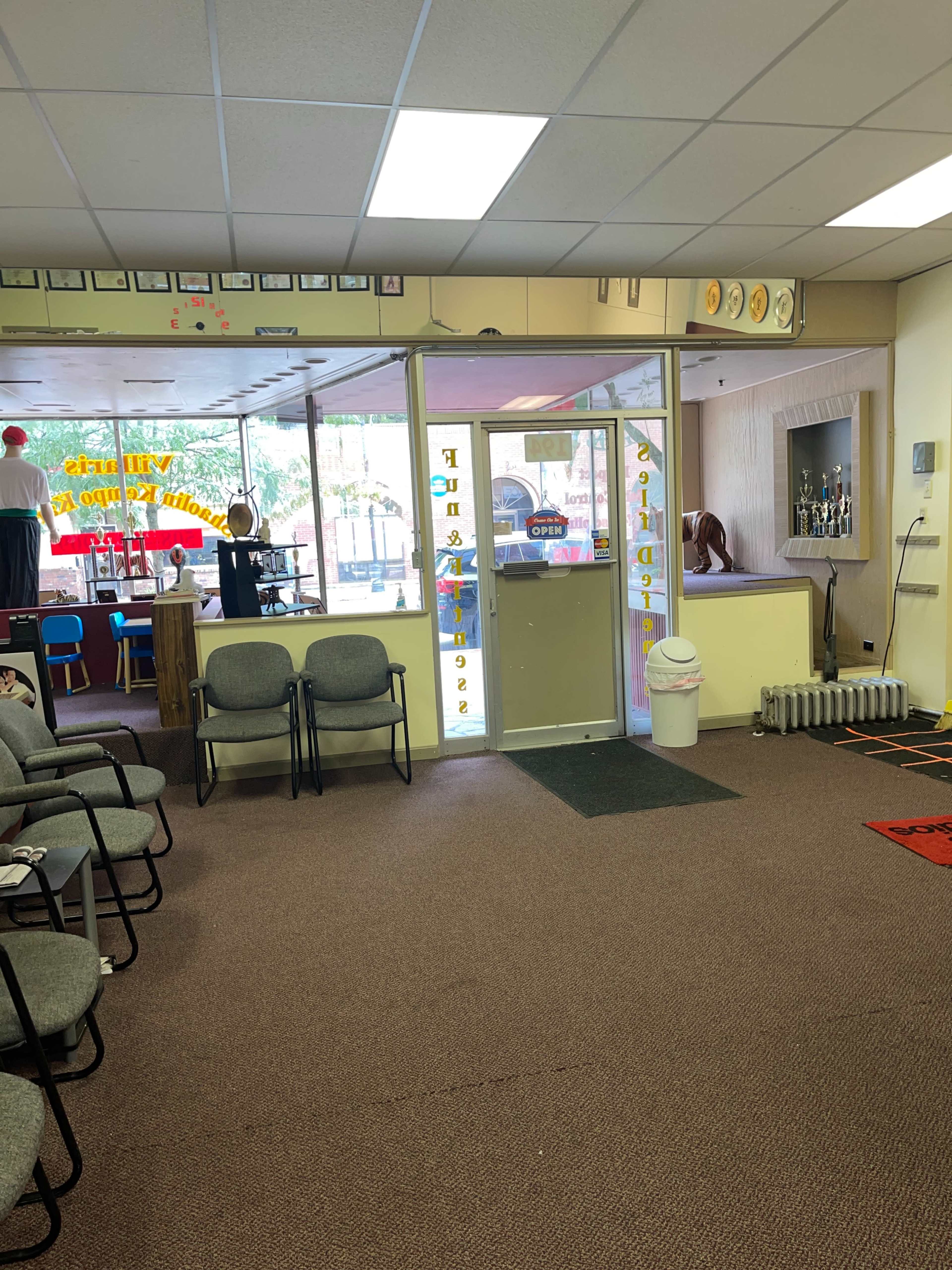 A waiting area with several chairs, a glass door leading to another room, and a visible reception area with trophies and decorations.