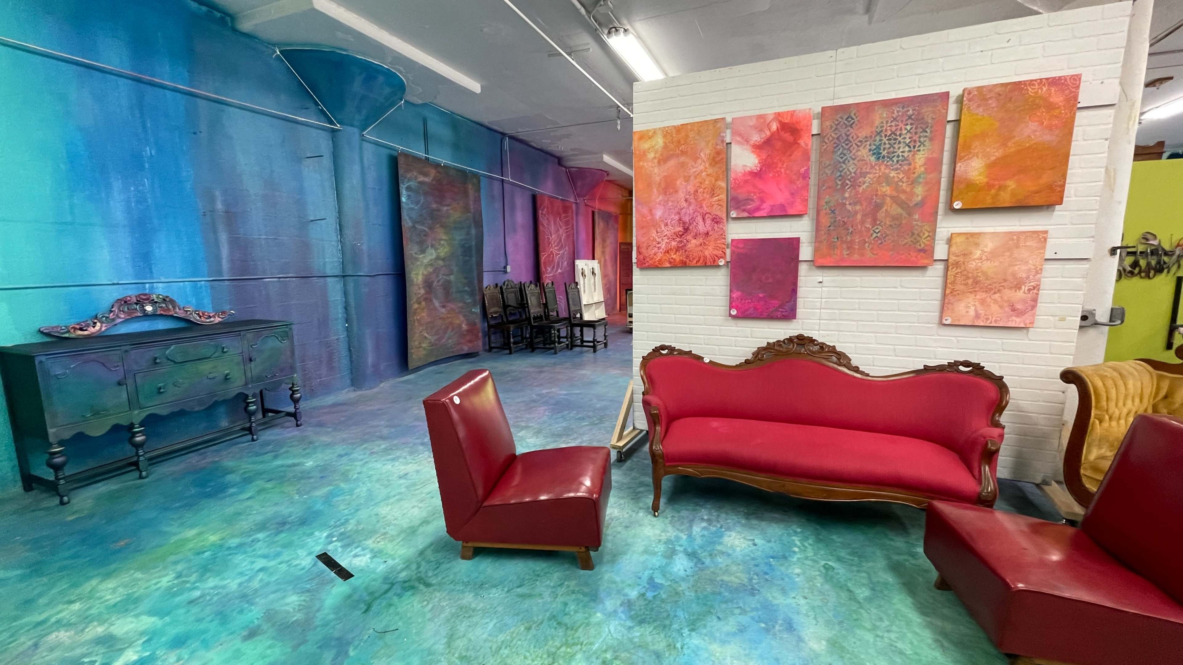 The image depicts a vibrant studio space with colorful walls and several artworks displayed alongside antique-style furniture, including a pink sofa and a red chair.