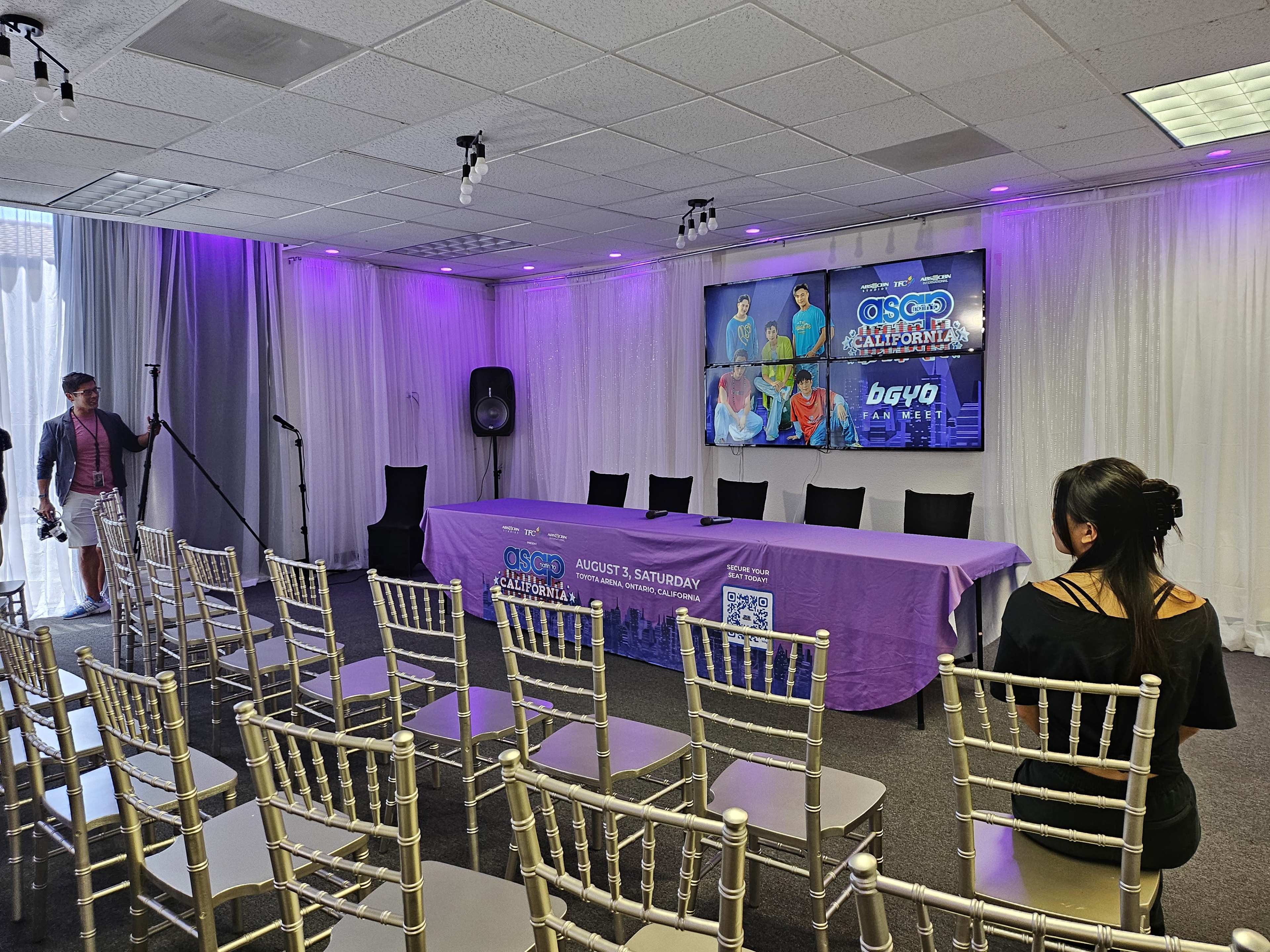 A conference room is set up with a purple tablecloth, empty chairs facing a stage with a screen displaying event graphics, and a microphone on a stand.