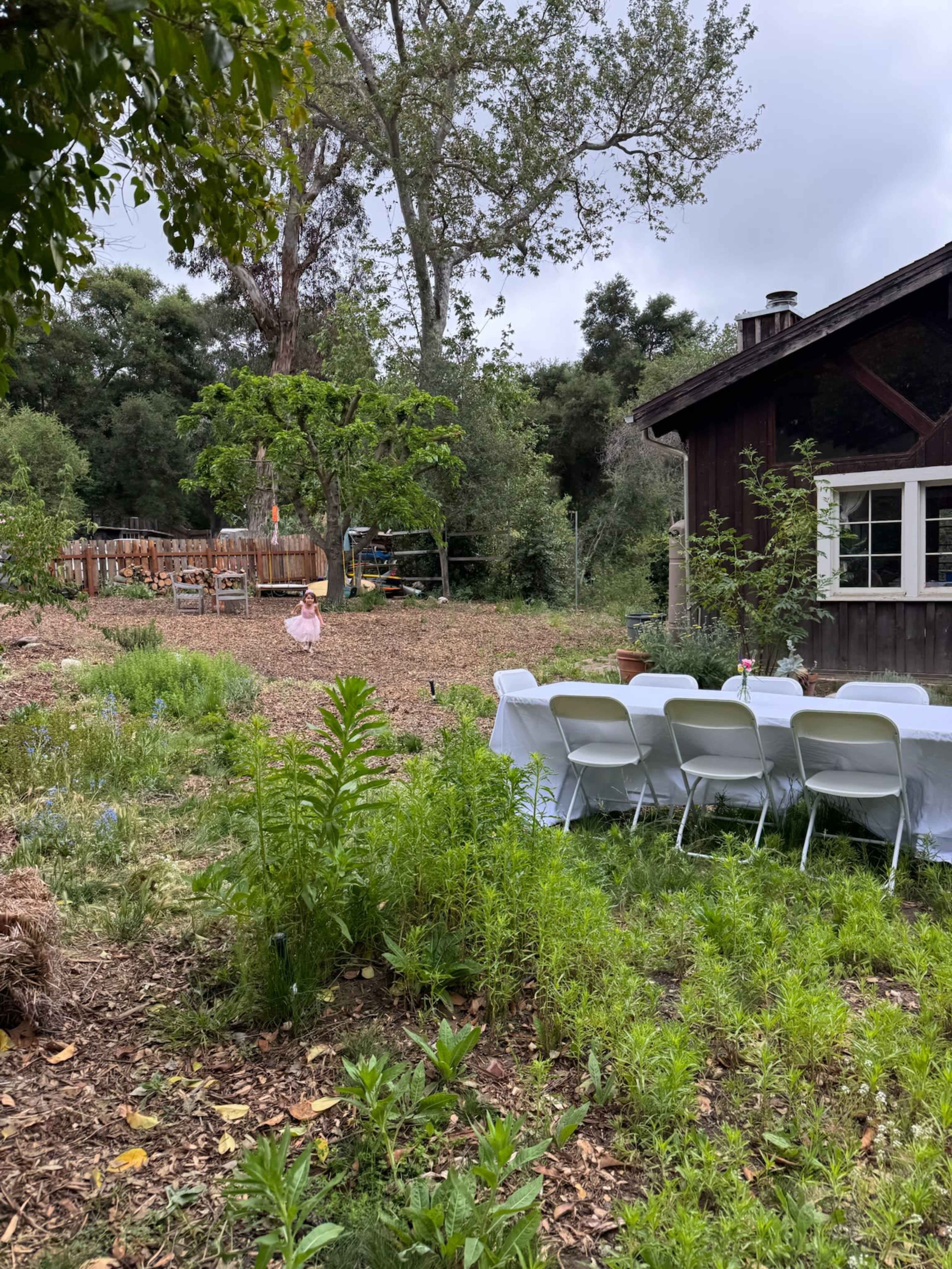 A long table with white chairs is set up in a garden area surrounded by greenery, with a rustic wooden building in the background.