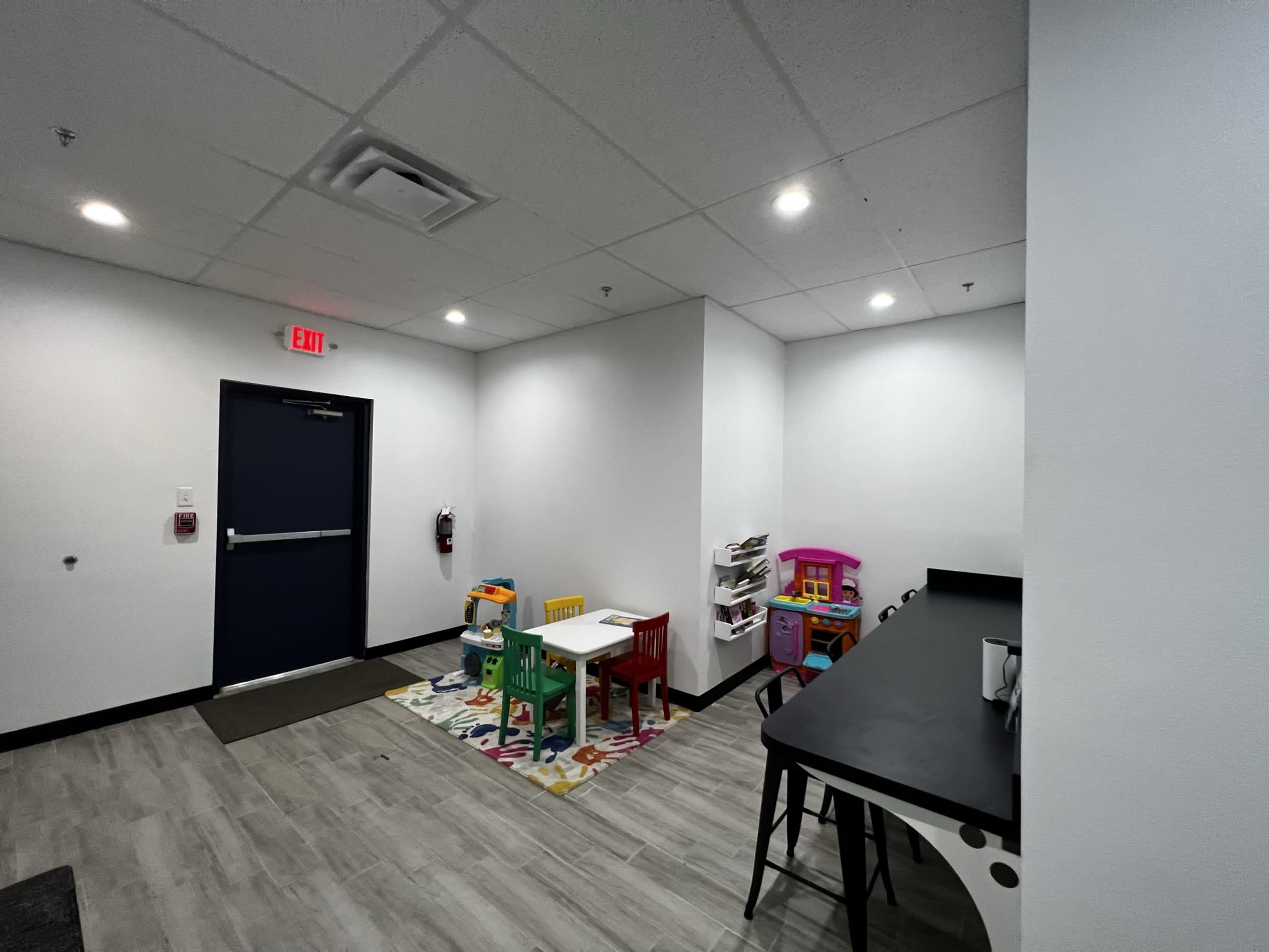 A brightly lit indoor play area featuring a small white table with colorful chairs, a toy kitchen set, and a playhouse on a patterned rug.