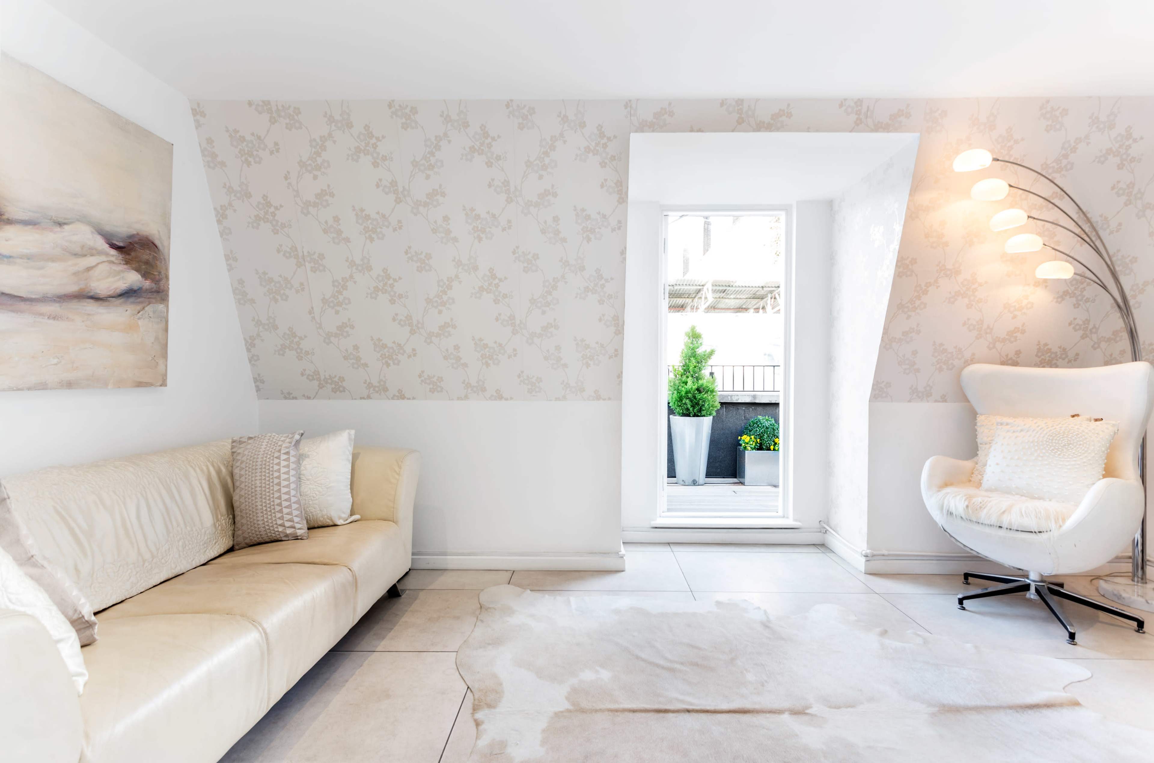 A modern living space with a light-colored sofa, a decorative rug, and a large window leading to a balcony with potted plants.
