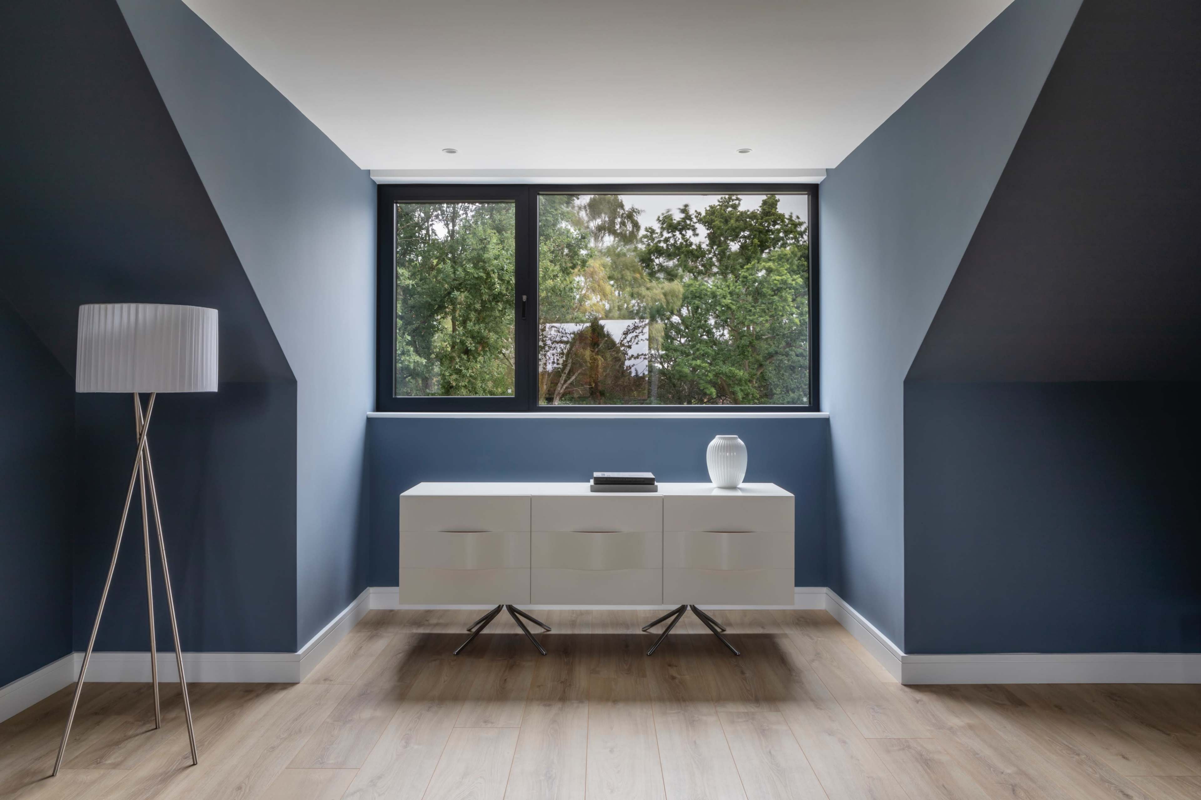 A modern room features a white sideboard and a tall lamp, with large windows providing a view of greenery outside.