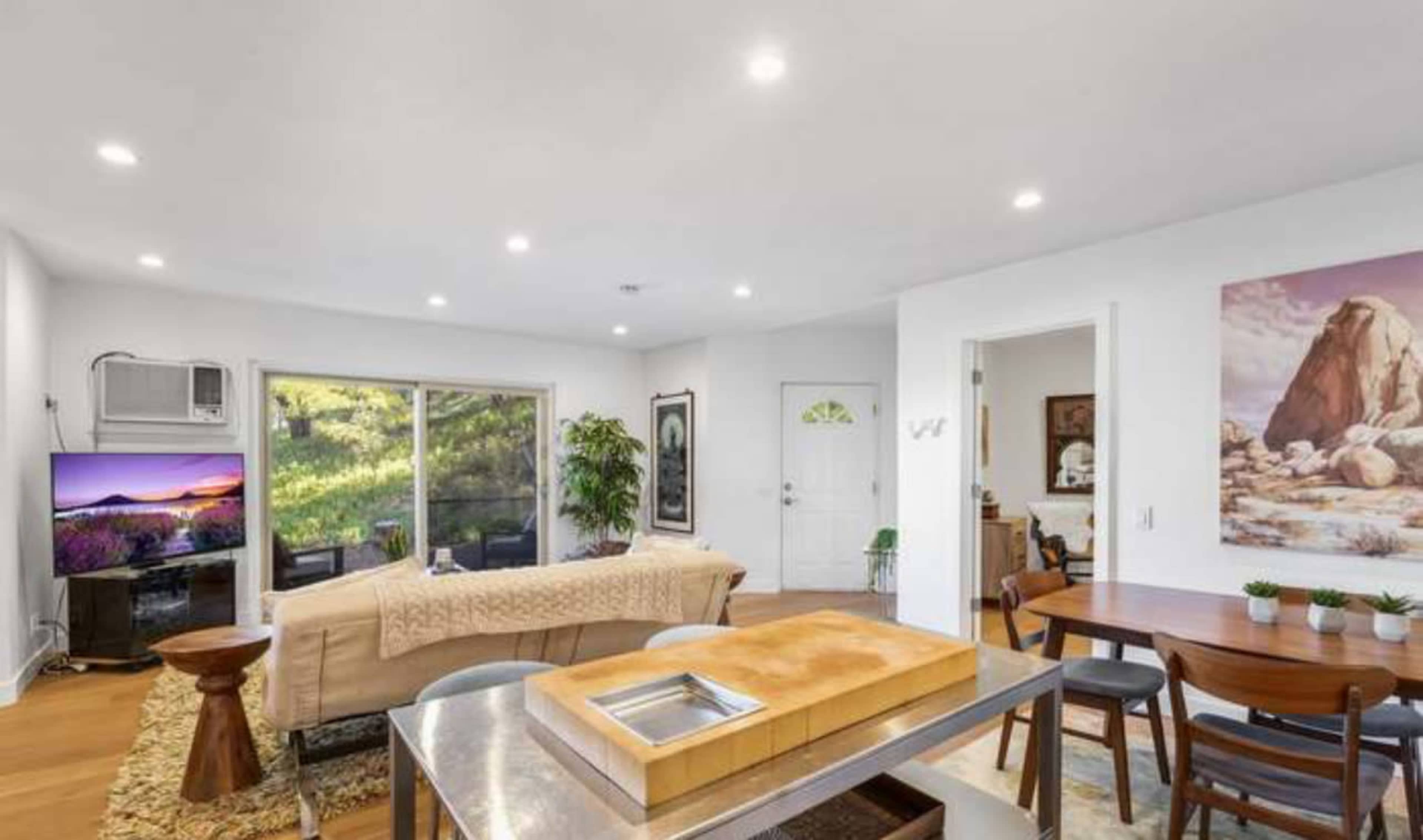 A bright living area features a sofa, a dining table, and a wall-mounted television, with large windows providing views of the outdoors.