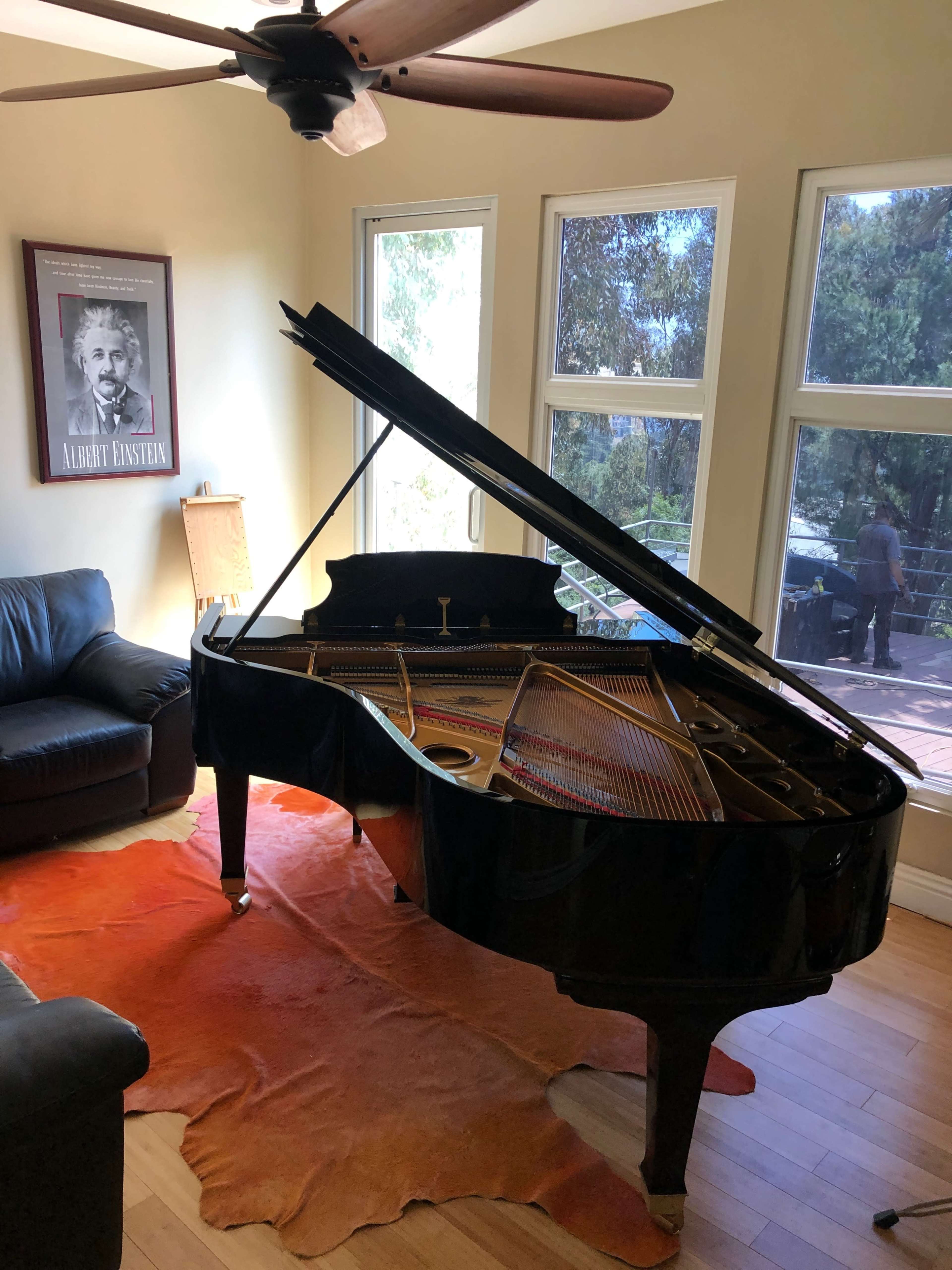 A grand piano with its lid open sits on a brown rug in a room with large windows, overlooking a patio area.