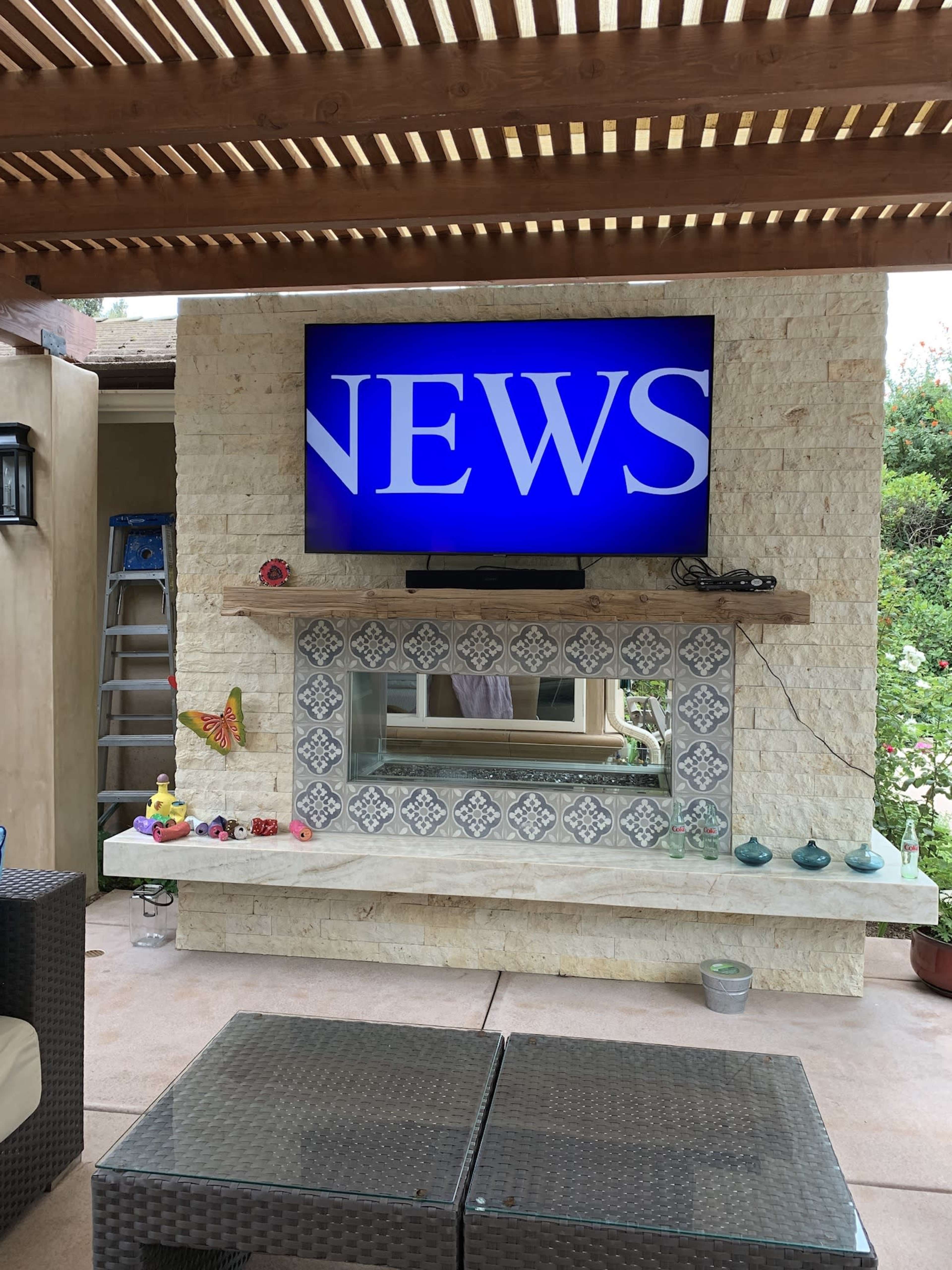 A television displaying the word "NEWS" is mounted on a stone wall above a decorative shelf in a patio area.
