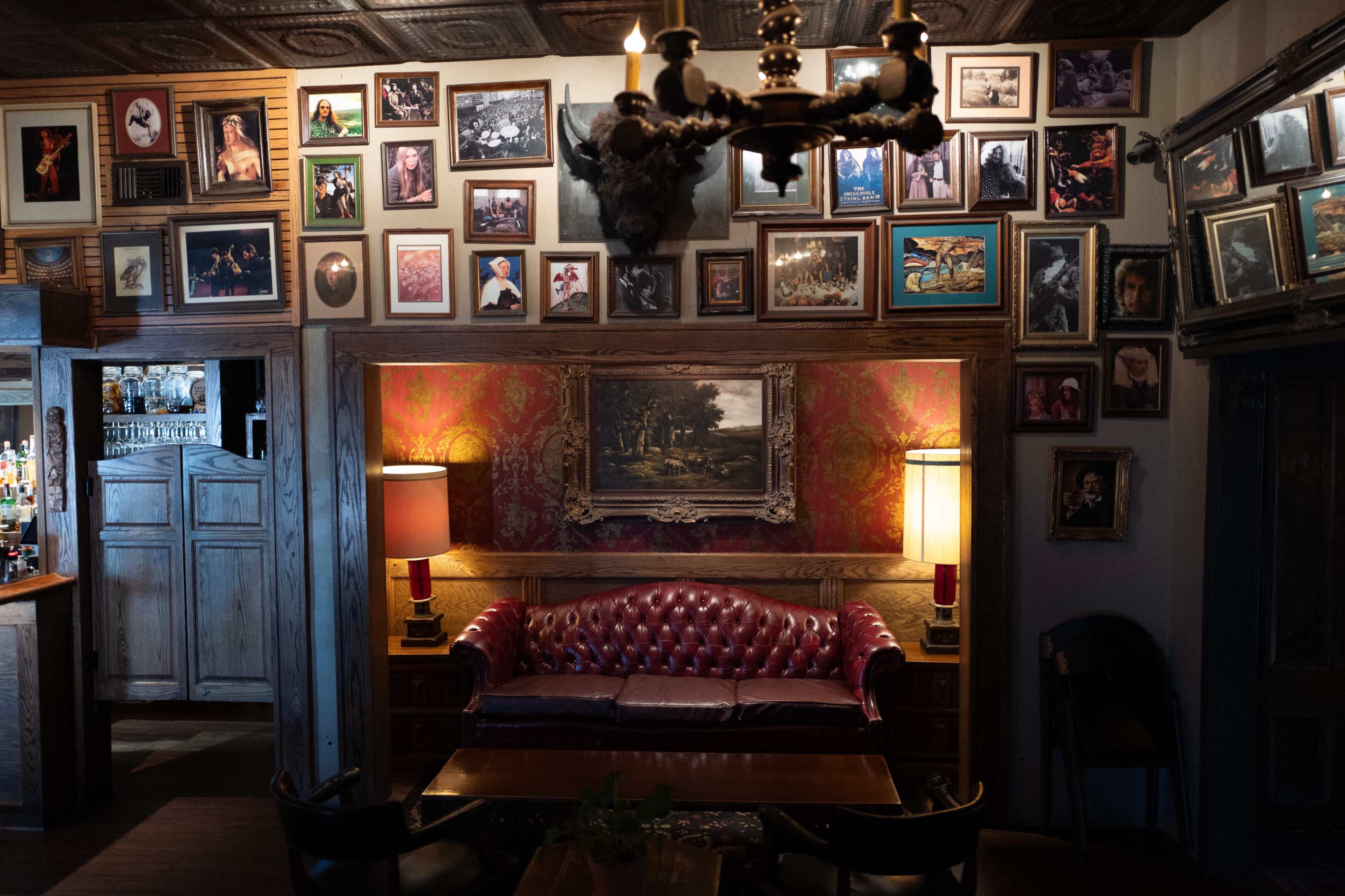 The image shows a cozy bar or lounge with a red tufted leather sofa, wooden tables, and walls adorned with various framed pictures and paintings.
