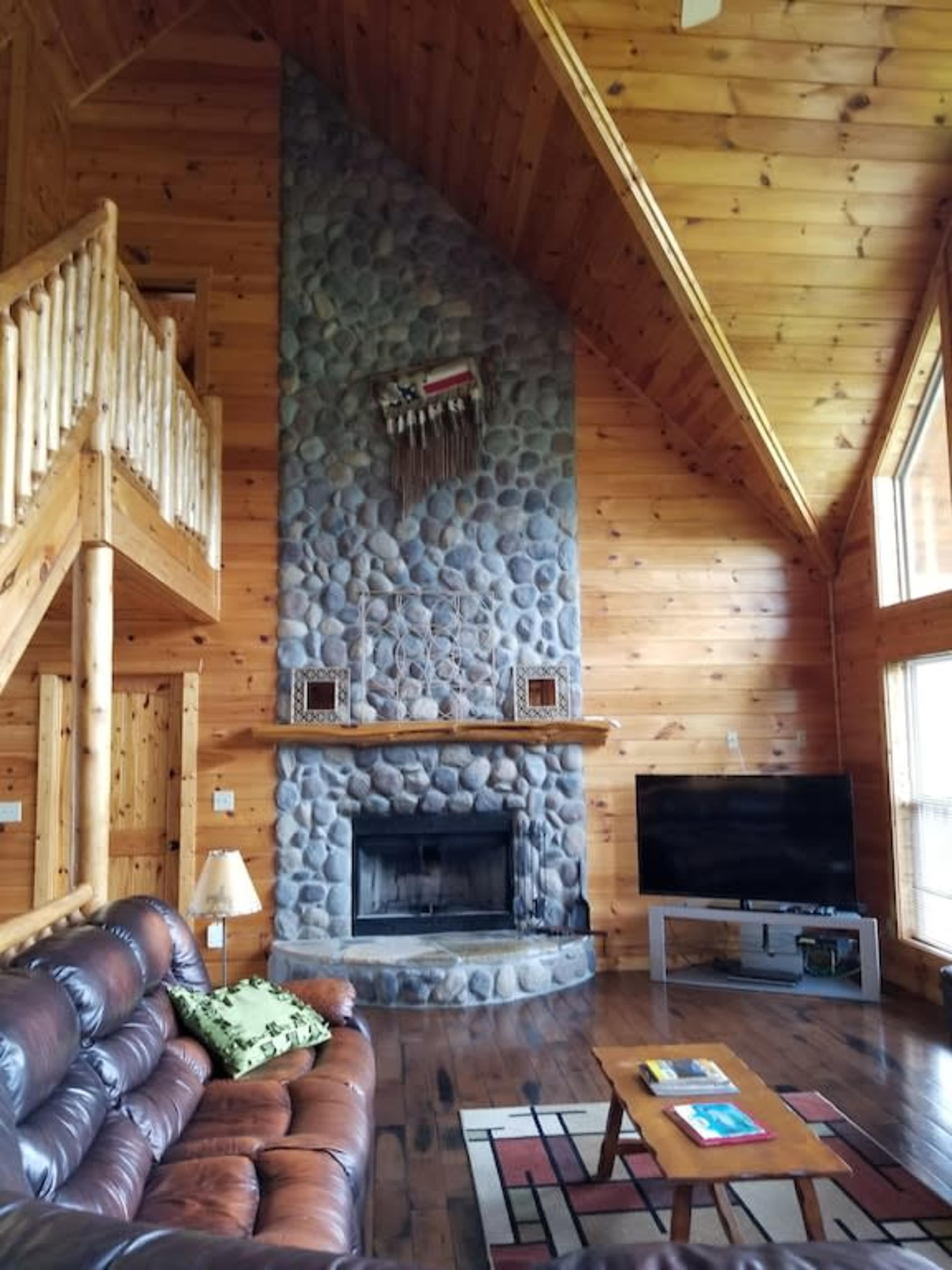 The interior of a wooden cabin features a stone fireplace, large windows, a leather sofa, and a staircase leading to an upper level.