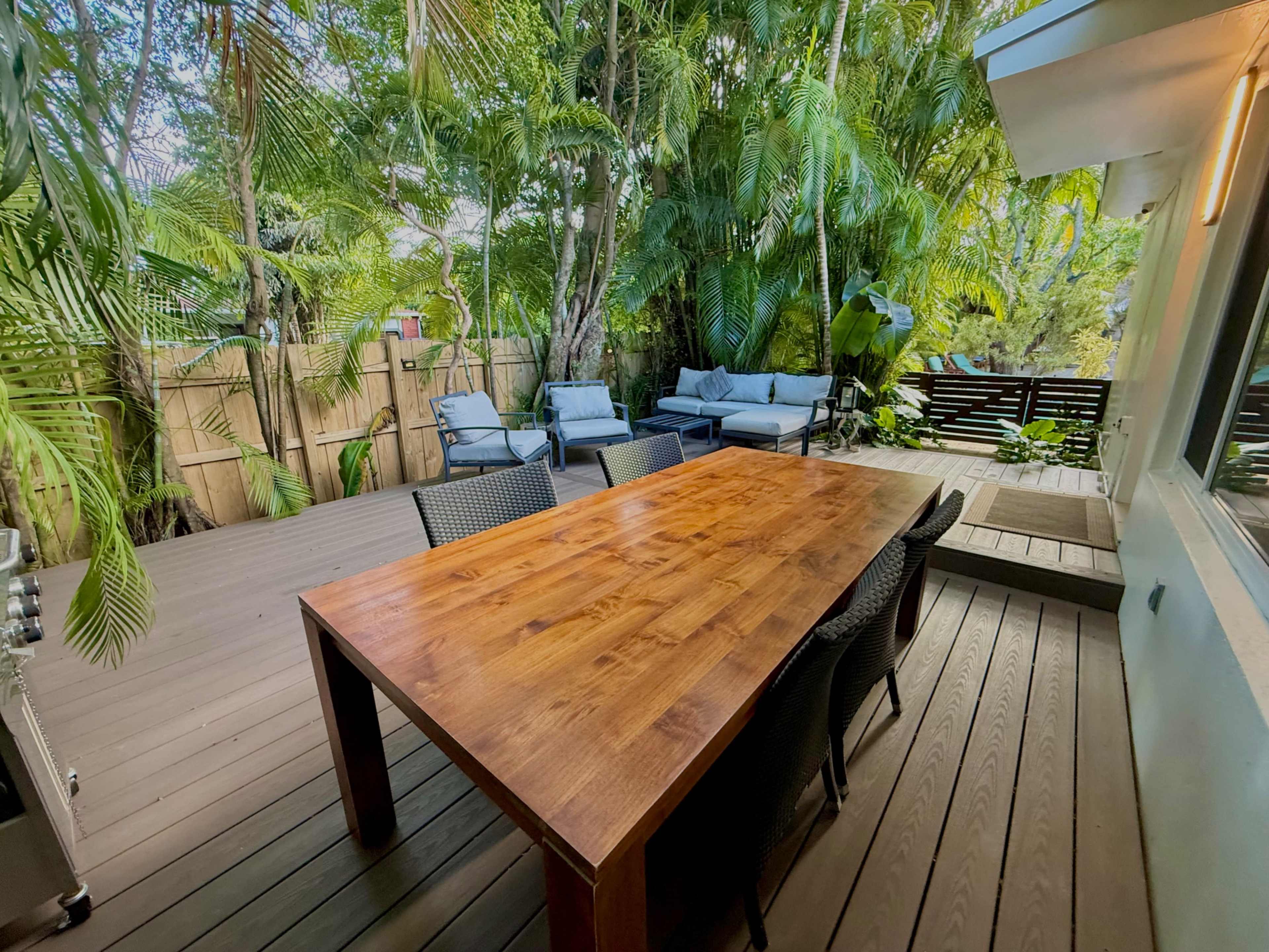 A wooden dining table with six chairs is positioned on a deck surrounded by lush green foliage and patio furniture.