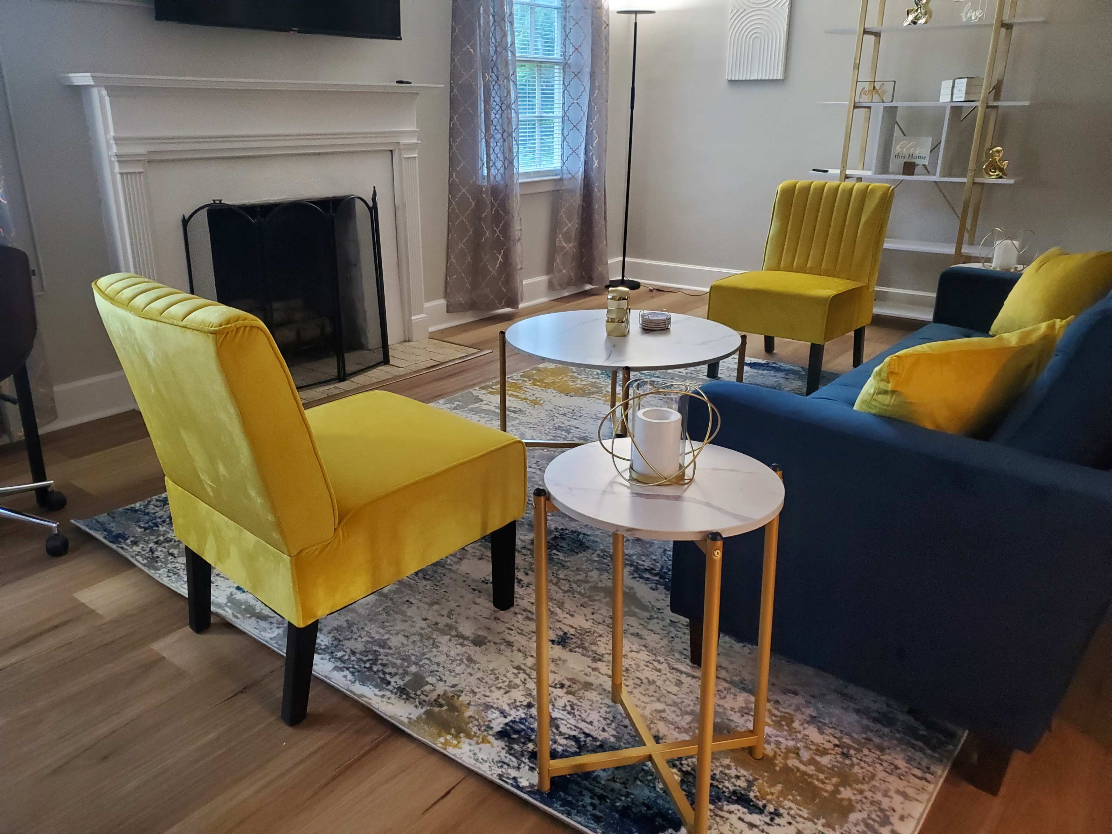 The image shows a modern living room with two yellow chairs and a blue sofa arranged around a small round table on a decorative rug, with a fireplace and a floor lamp in the background.