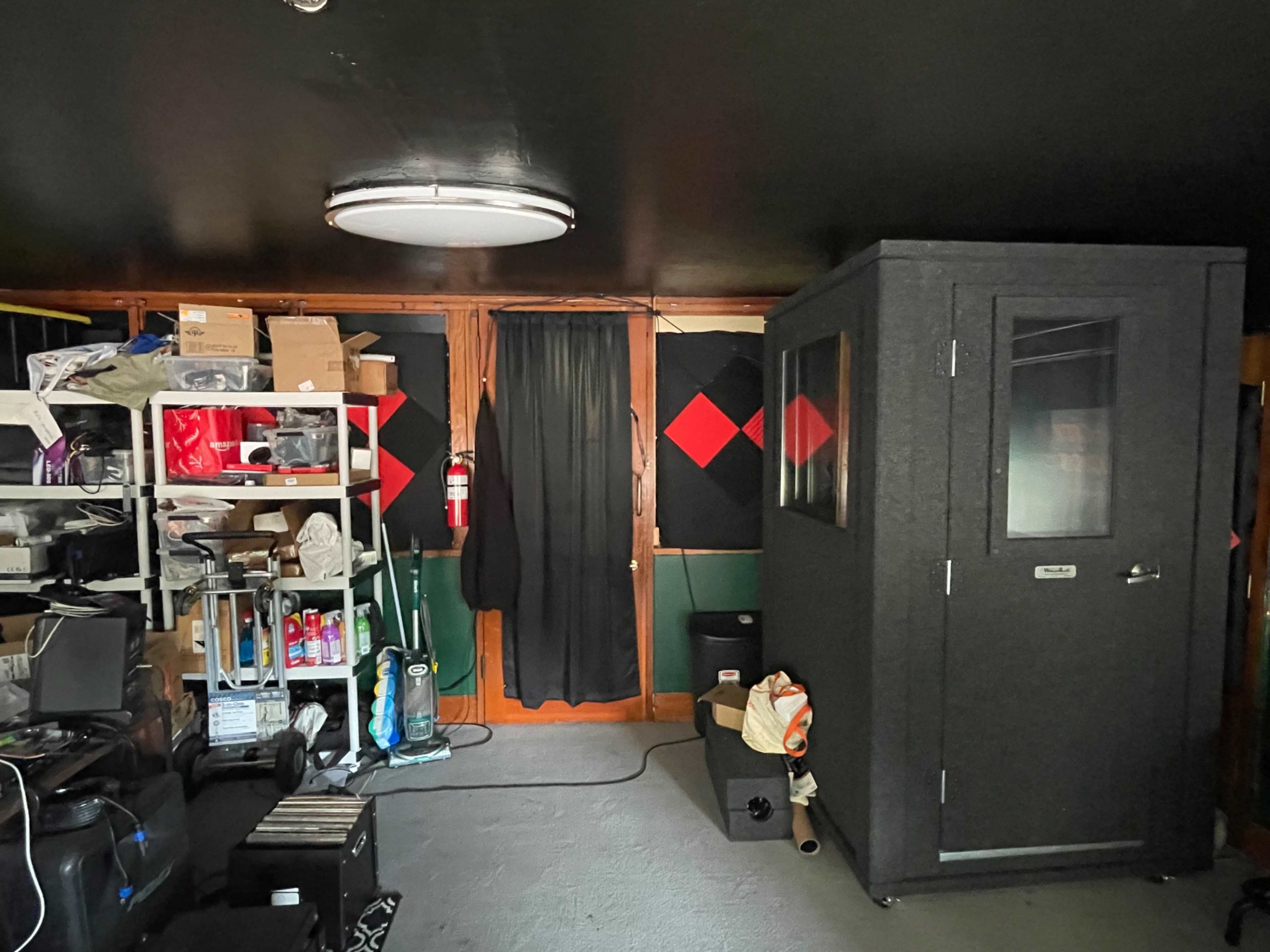 A cluttered room with shelves full of boxes and equipment, a black sound booth, and a door covered by a curtain in the background.