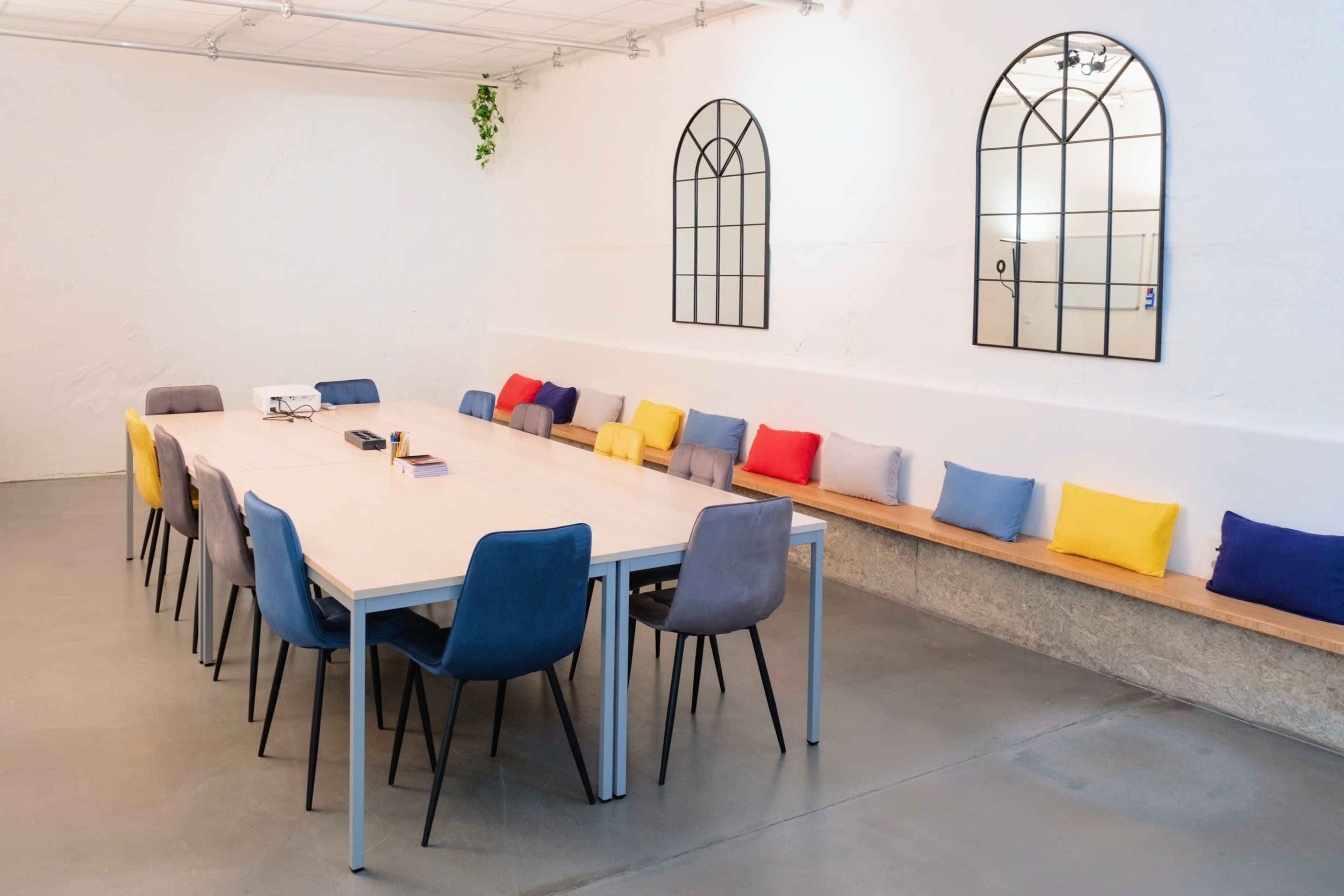 A long table surrounded by colorful chairs and a bench with cushions is set in a bright, minimalist meeting room.