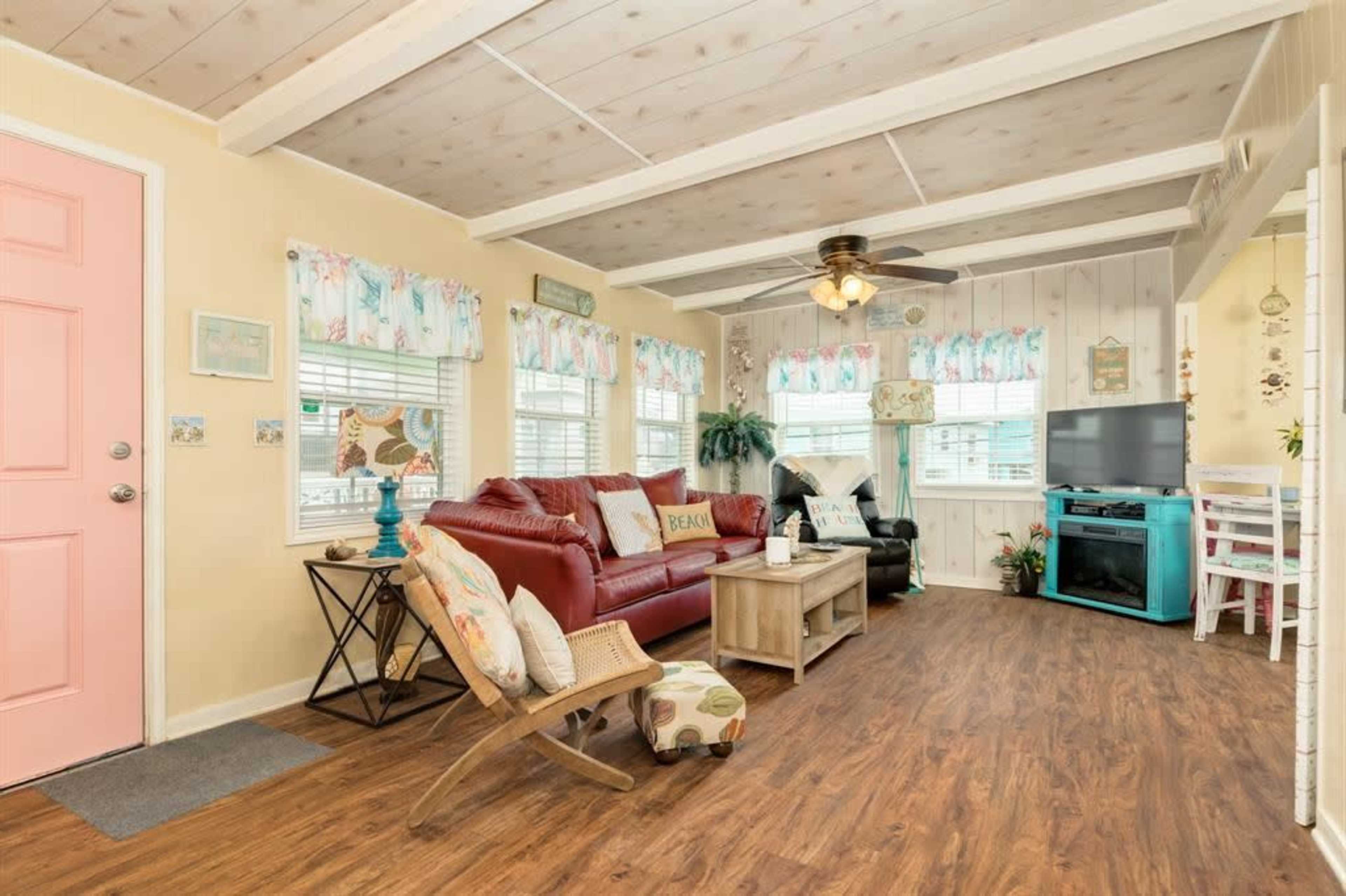 The image shows a living room with a red sofa, a light wooden coffee table, and a turquoise TV cabinet, surrounded by large windows with patterned curtains.