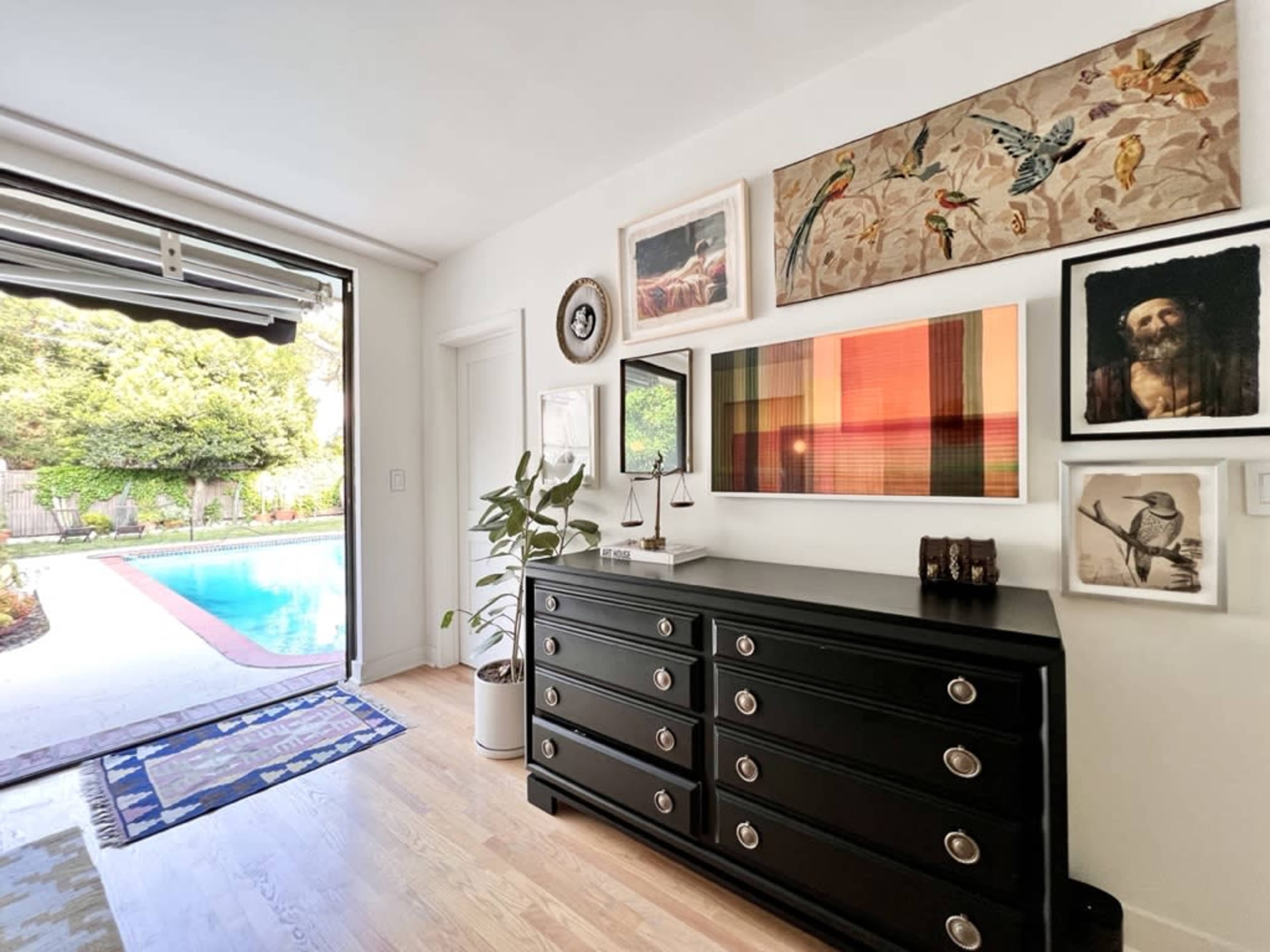 A modern room features a black dresser against a wall adorned with various framed artworks, with a view of a pool area through an open sliding door.