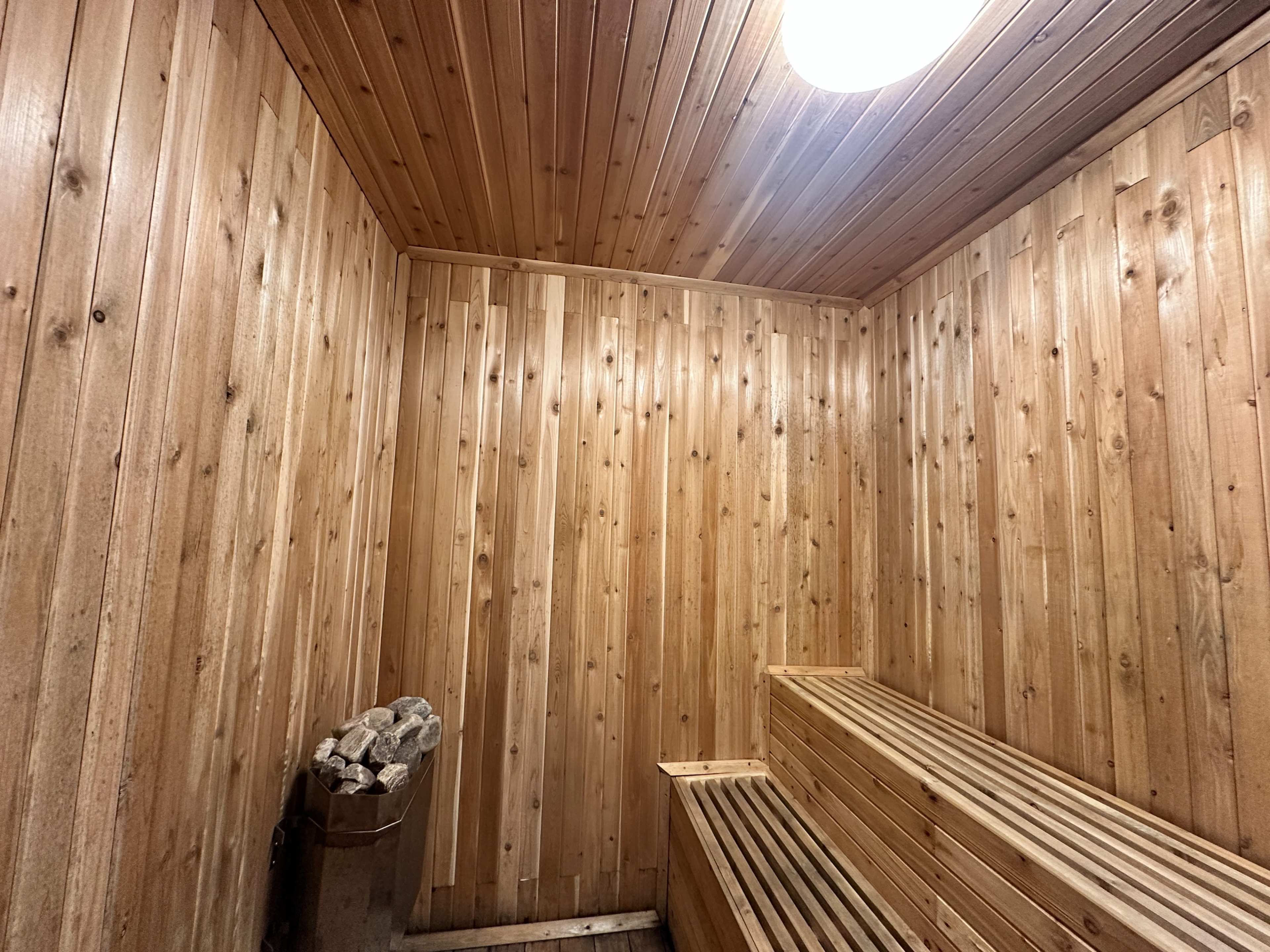 The image shows the interior of a wooden sauna with paneled walls, a bench along one side, and a stove with stacked stones nearby.