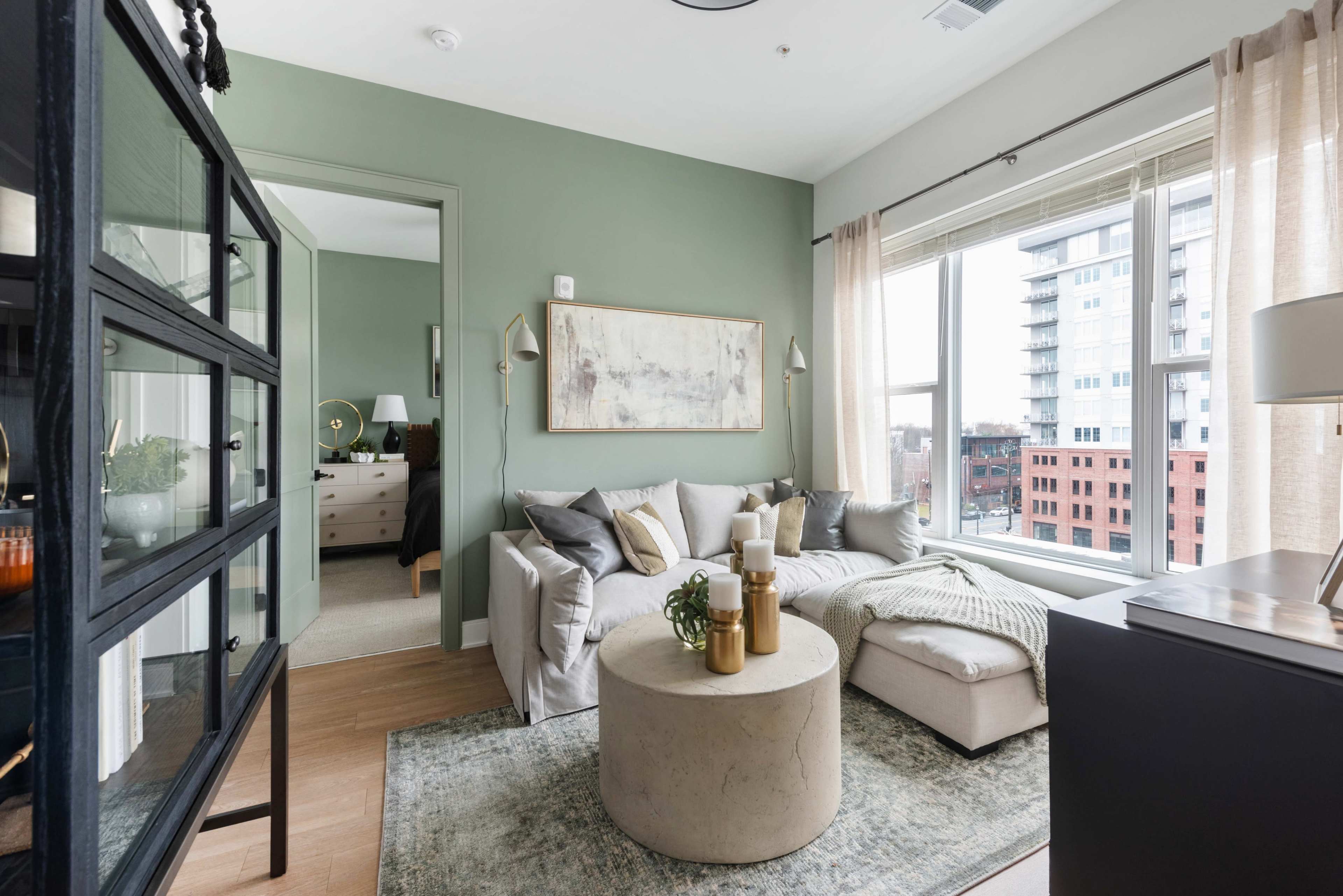 The image shows a cozy living area with a light-colored sofa, a round coffee table, and large windows that let in natural light, framed by sheer curtains.