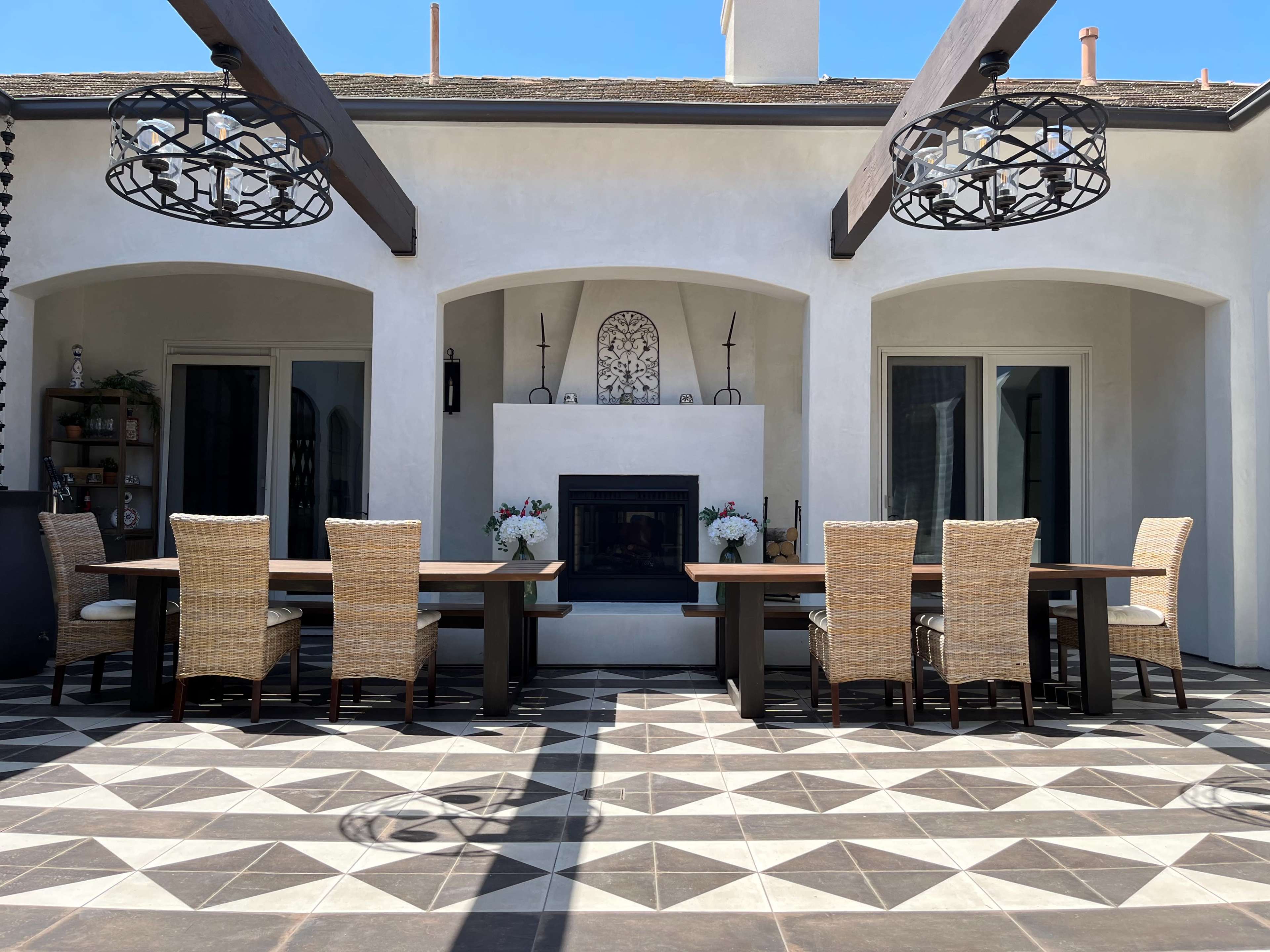 The image shows an outdoor dining area with two long tables surrounded by wicker chairs, a fireplace in the center, and large arching doorways.