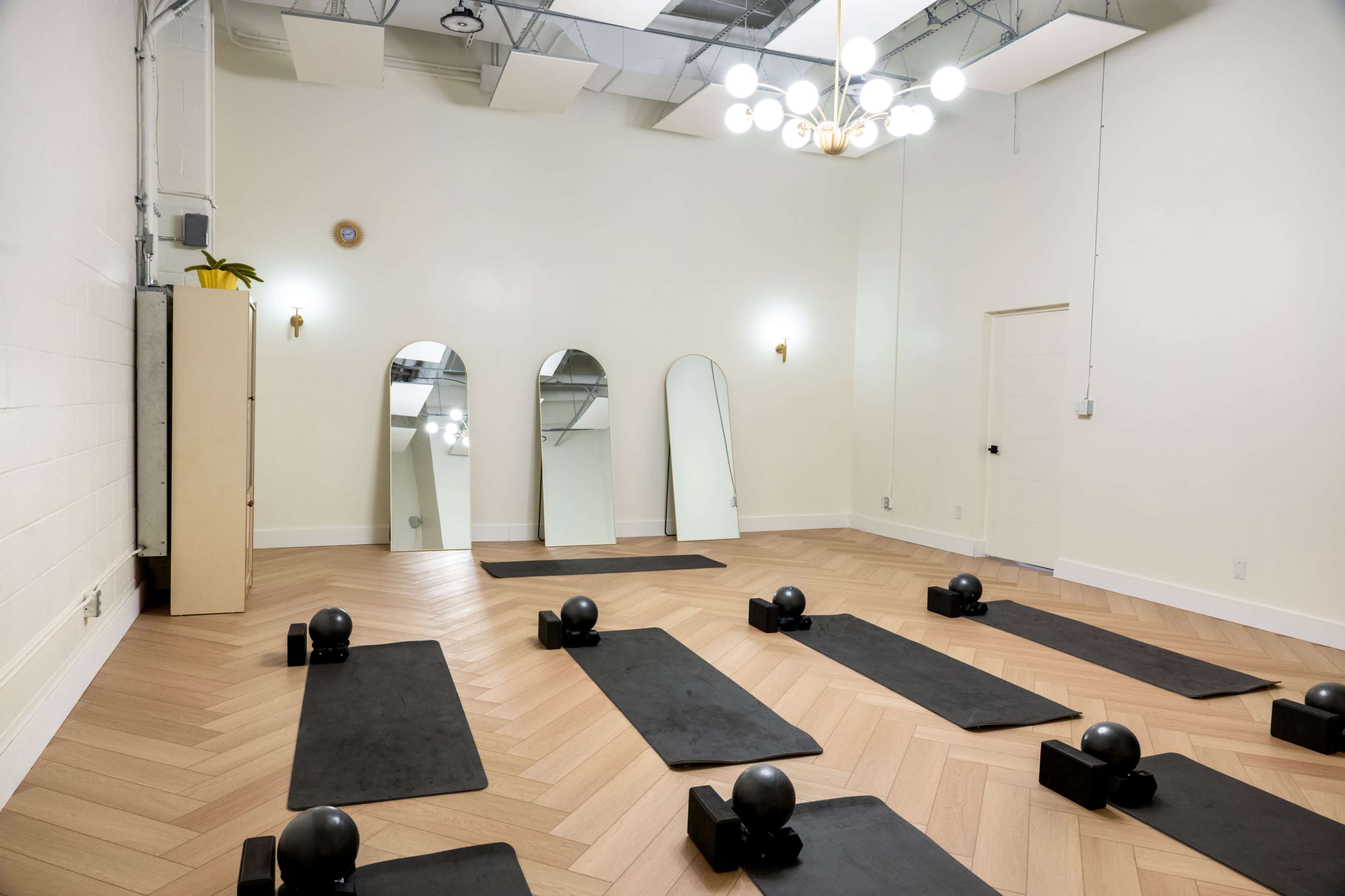 A bright, empty fitness studio features wooden flooring, exercise mats arranged on the ground, and mirrors hanging on the wall.