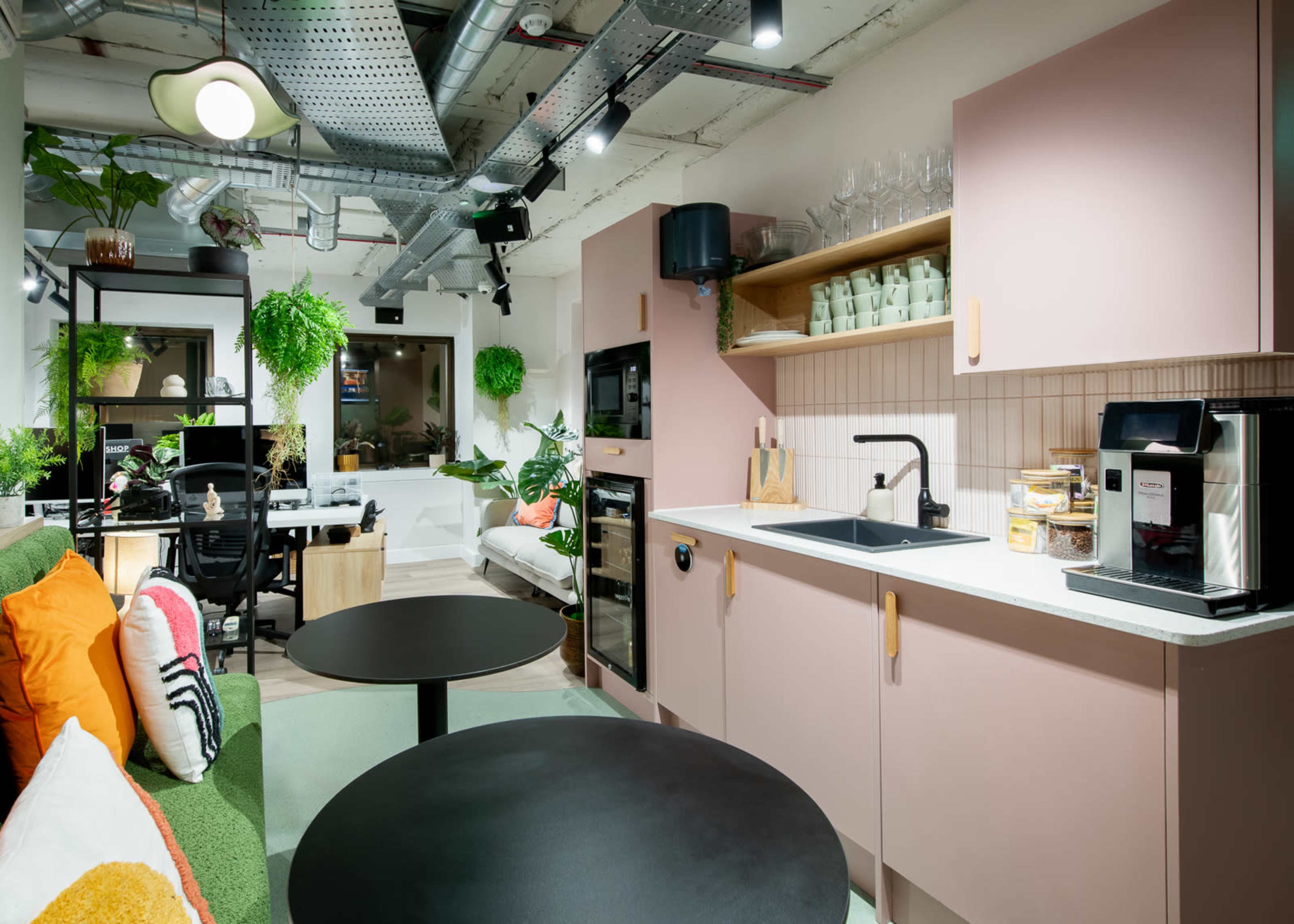 A modern kitchen area features pink cabinets, a black sink, and a coffee maker, with green plants and a seating area in the background.