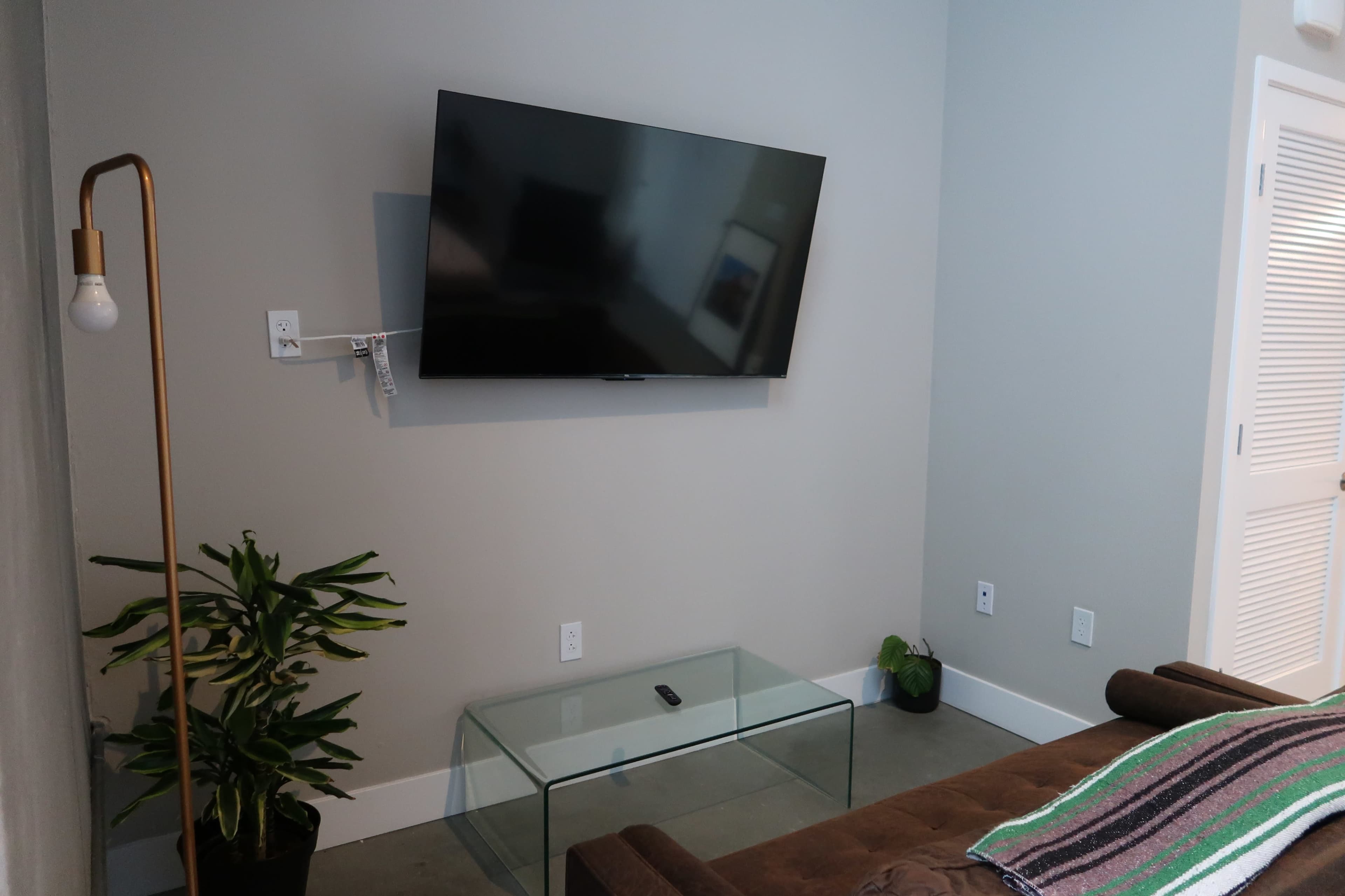 A modern living room features a wall-mounted television, a glass coffee table, a floor lamp, and a potted plant.