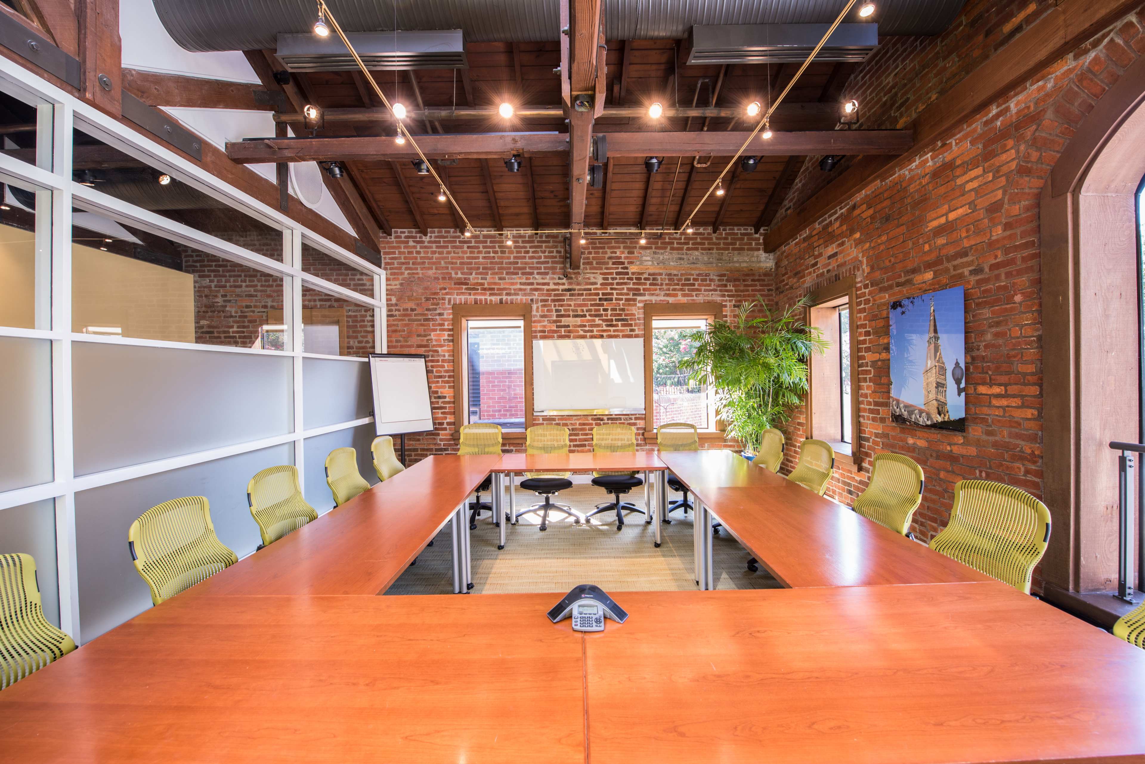 A spacious conference room features a large wooden table surrounded by yellow office chairs and a presentation board against a brick wall backdrop.