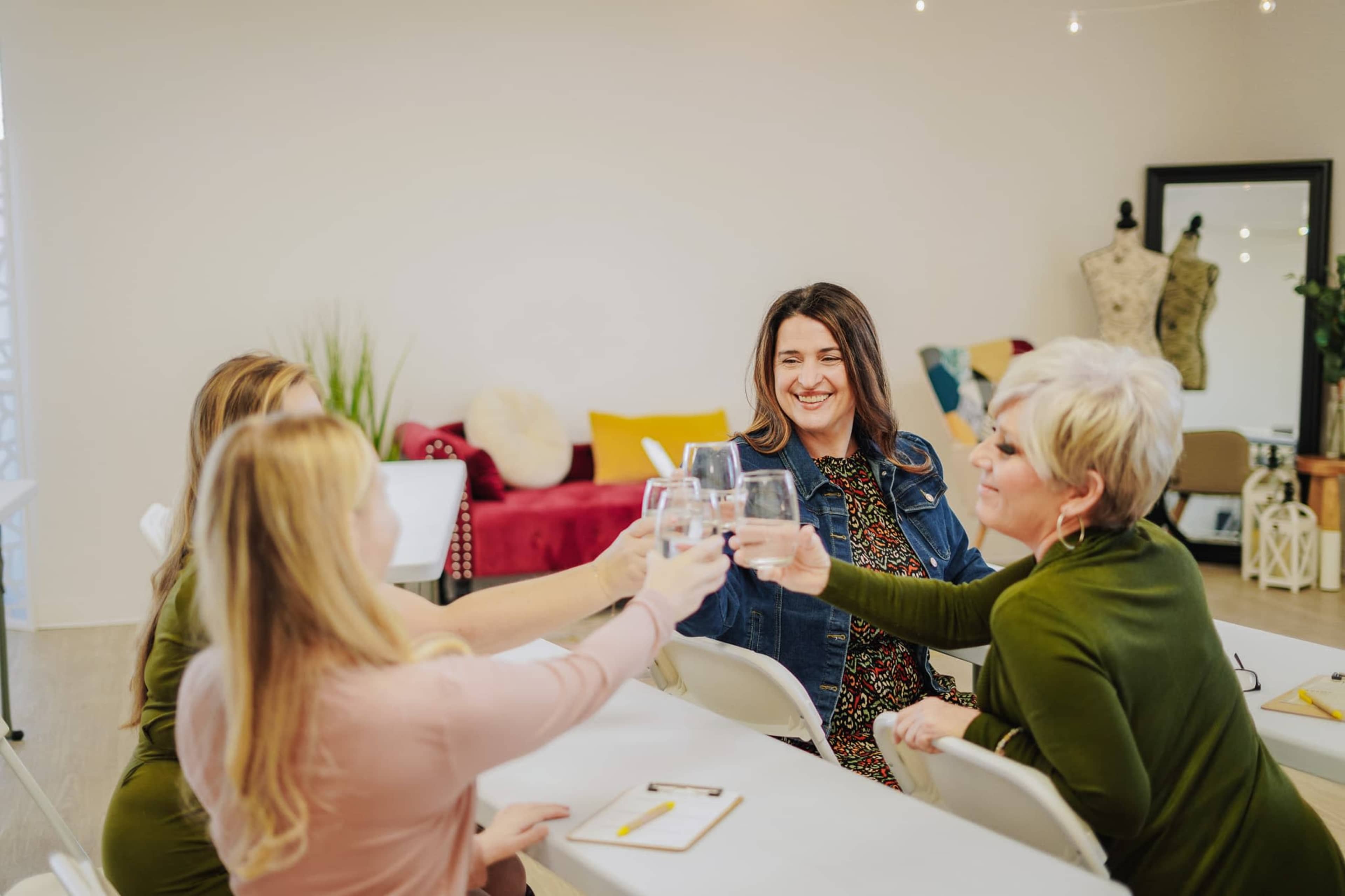 A group of five women is gathered around a table, smiling and toasting with glasses in a brightly lit room.