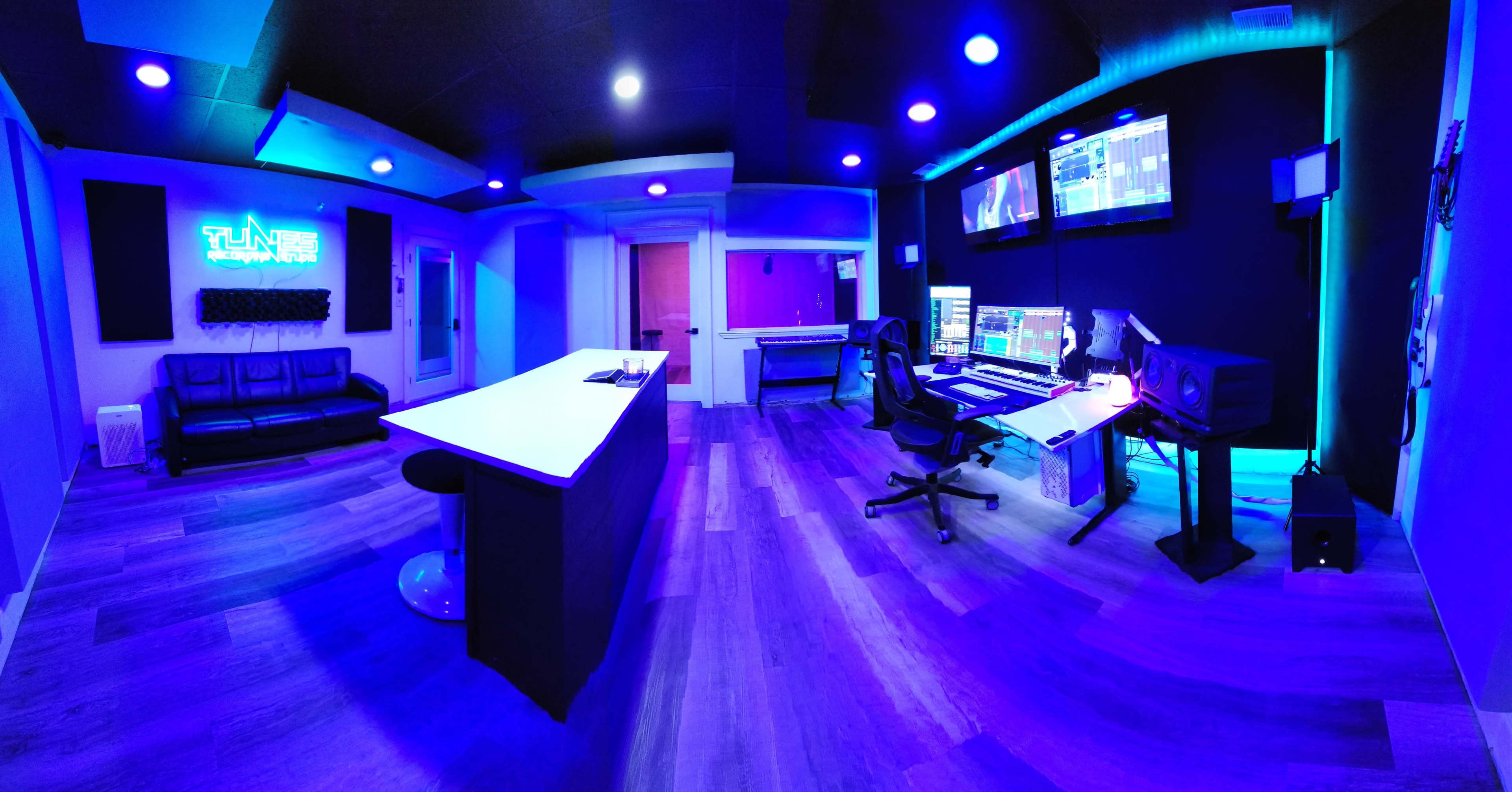 A modern recording studio with a long counter, a large desk equipped with audio equipment, and blue ambient lighting.