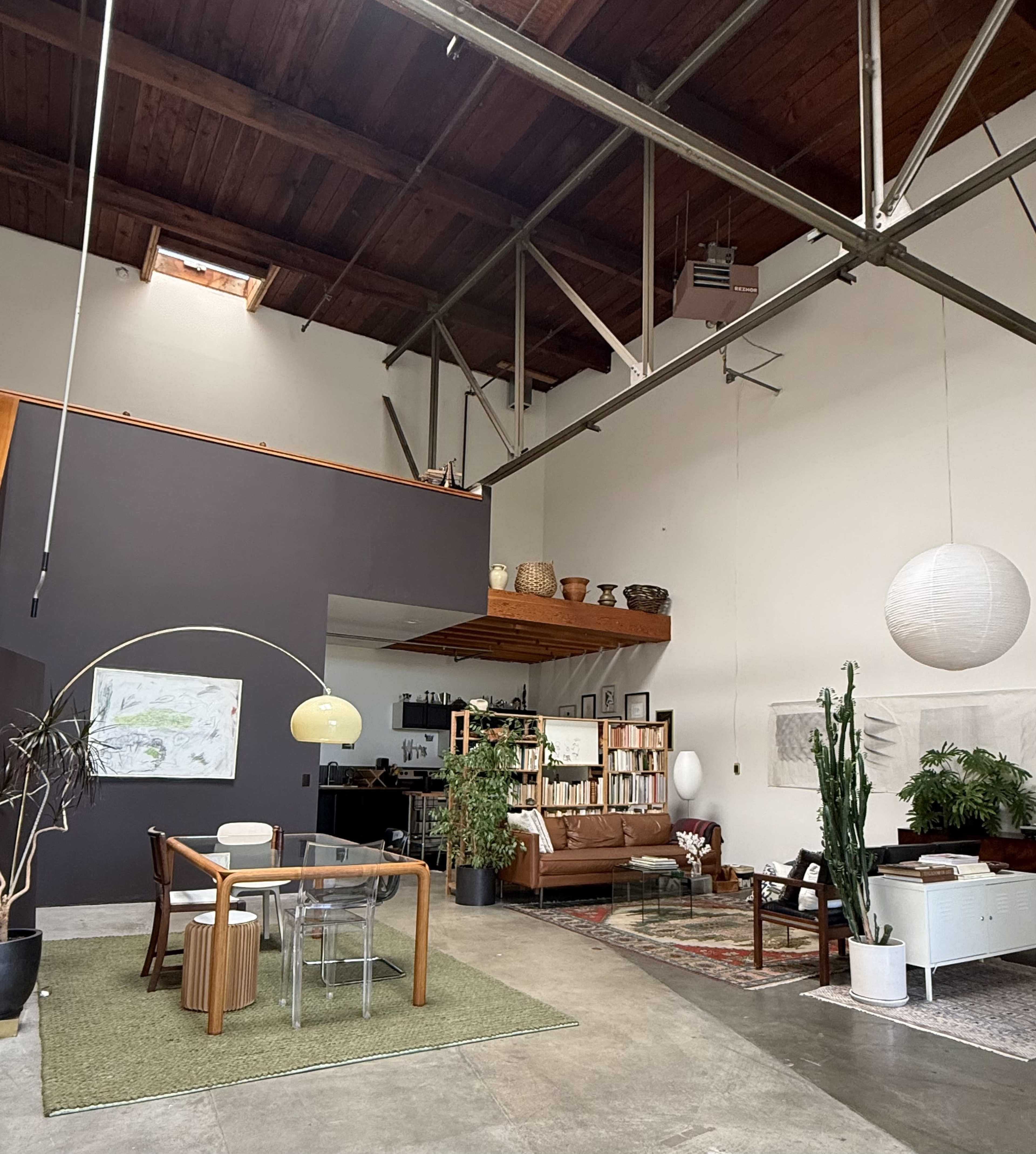A spacious, modern interior features a dining area with a glass table and wooden chairs, a comfortable seating area with a sofa, and large plants, all under a high ceiling with exposed beams.