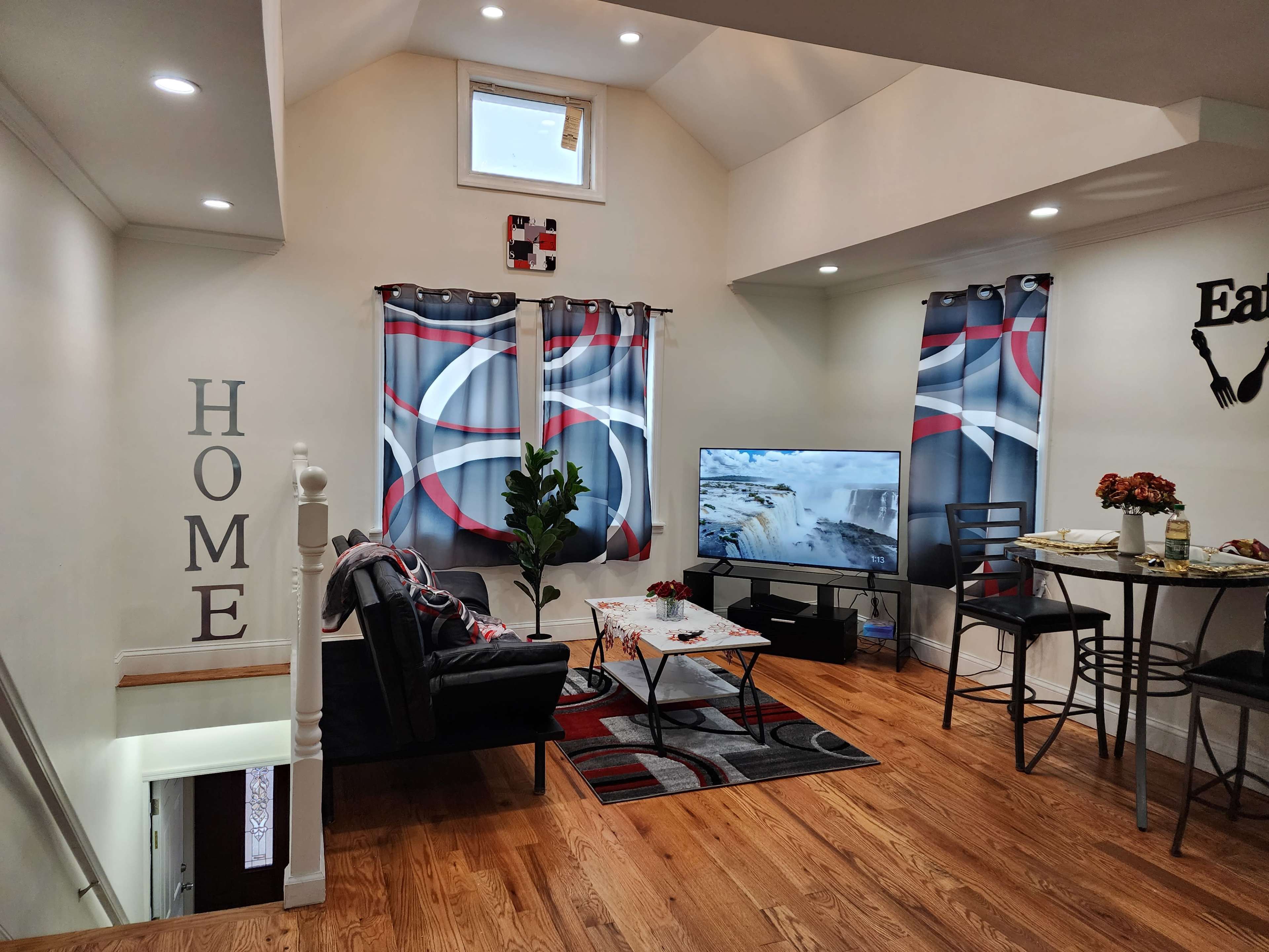 The image shows a modern living room with a black sofa, a coffee table adorned with decorative items, and a television displaying a nature scene, all set beneath a high ceiling with large windows.