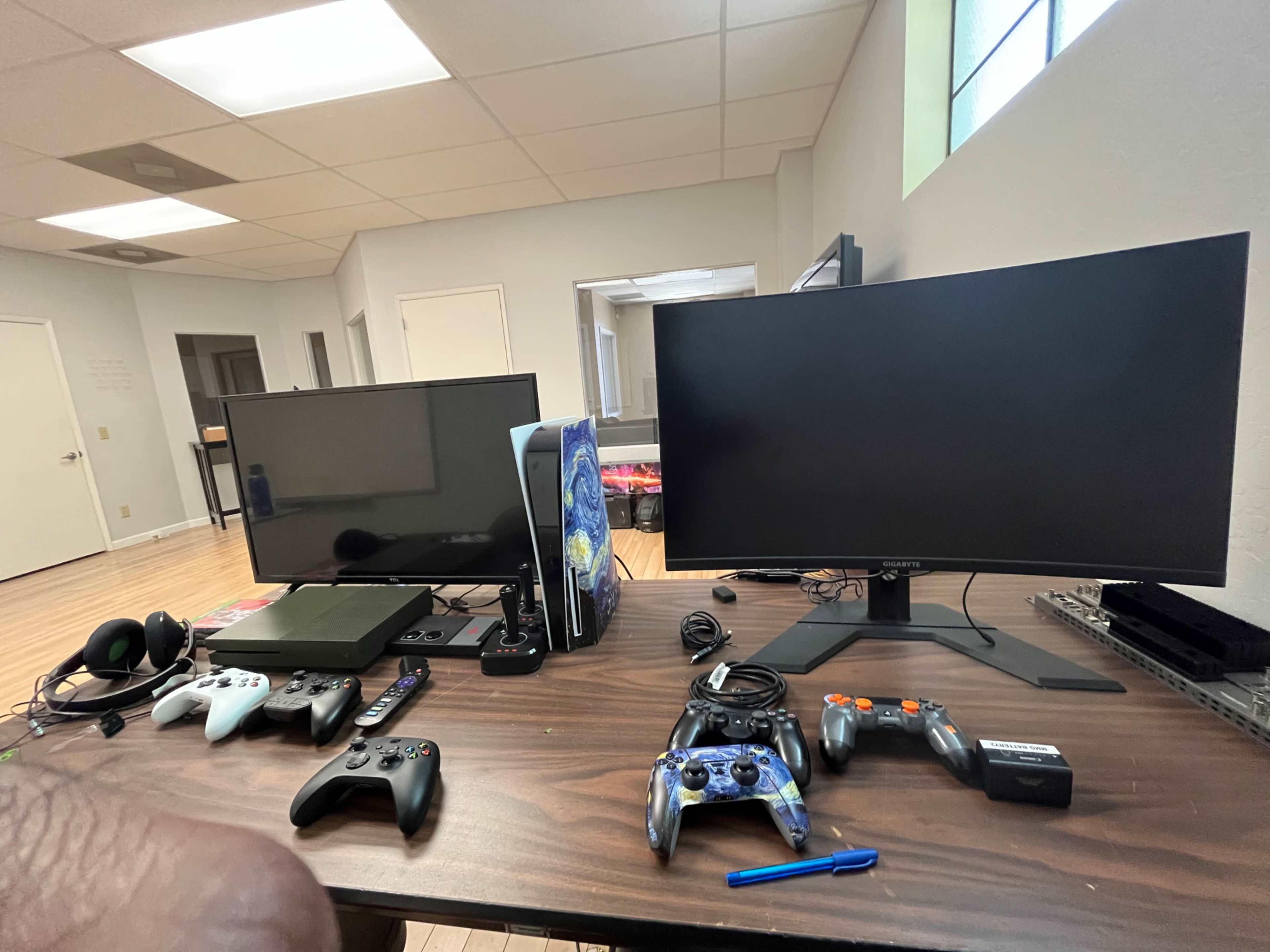 The image shows a table with two monitors, several gaming consoles, and multiple game controllers arranged on it.