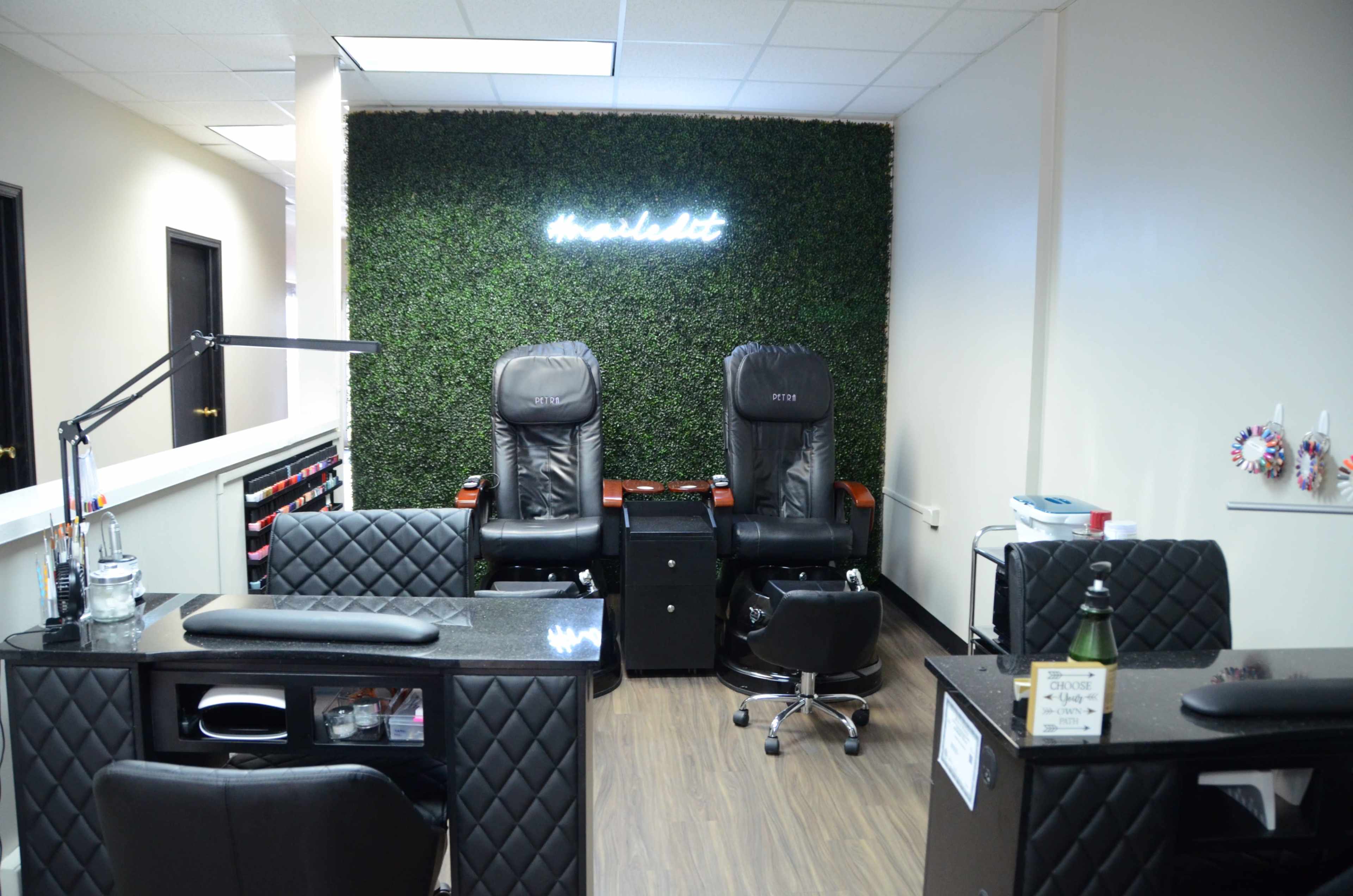 The image shows a nail salon with two black manicure chairs, a reception desk, and a greenery wall featuring illuminated signage.