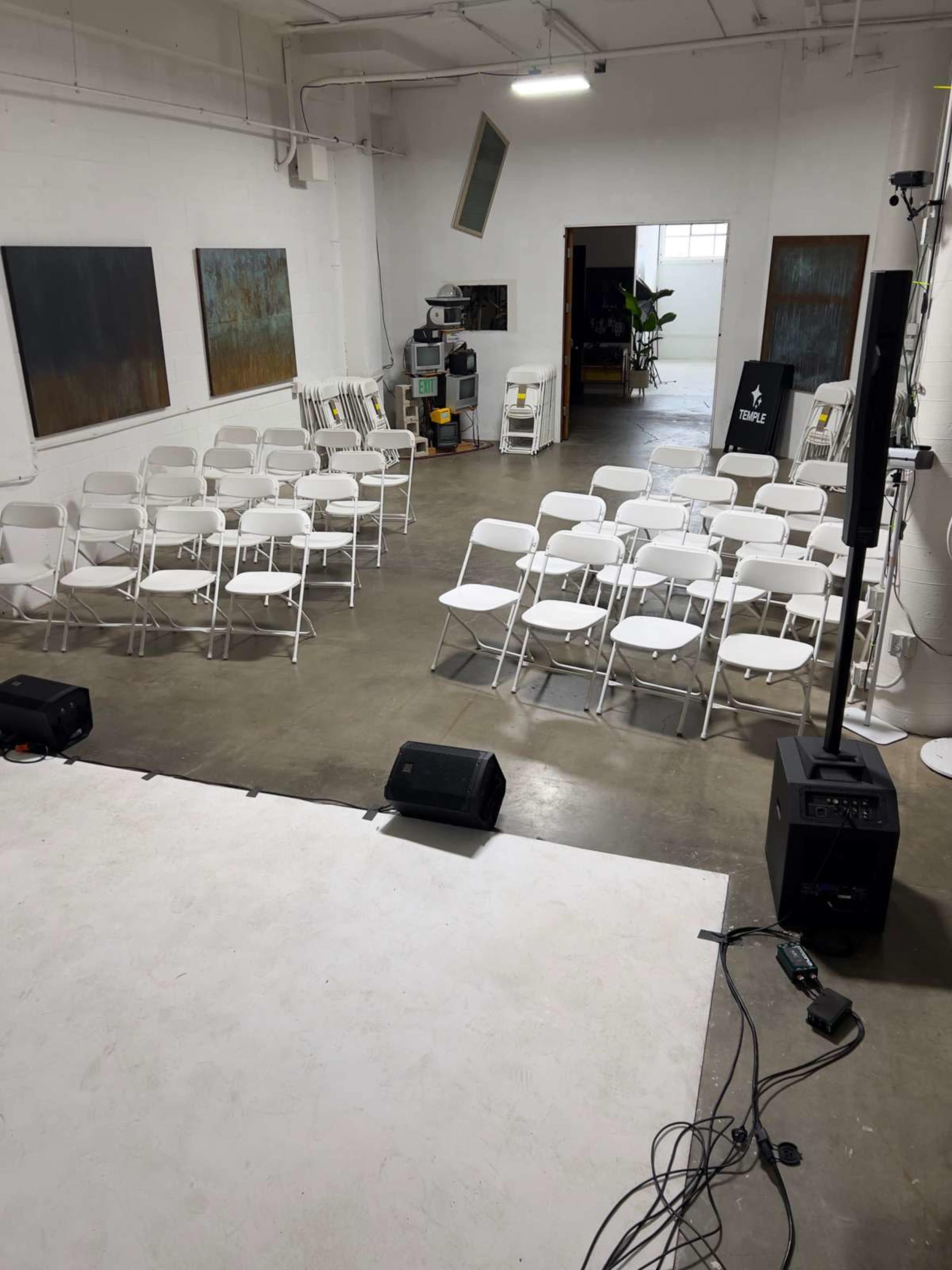 The image shows a spacious indoor venue featuring rows of white folding chairs arranged for an event, with a small elevated stage and various audio equipment visible in the foreground.