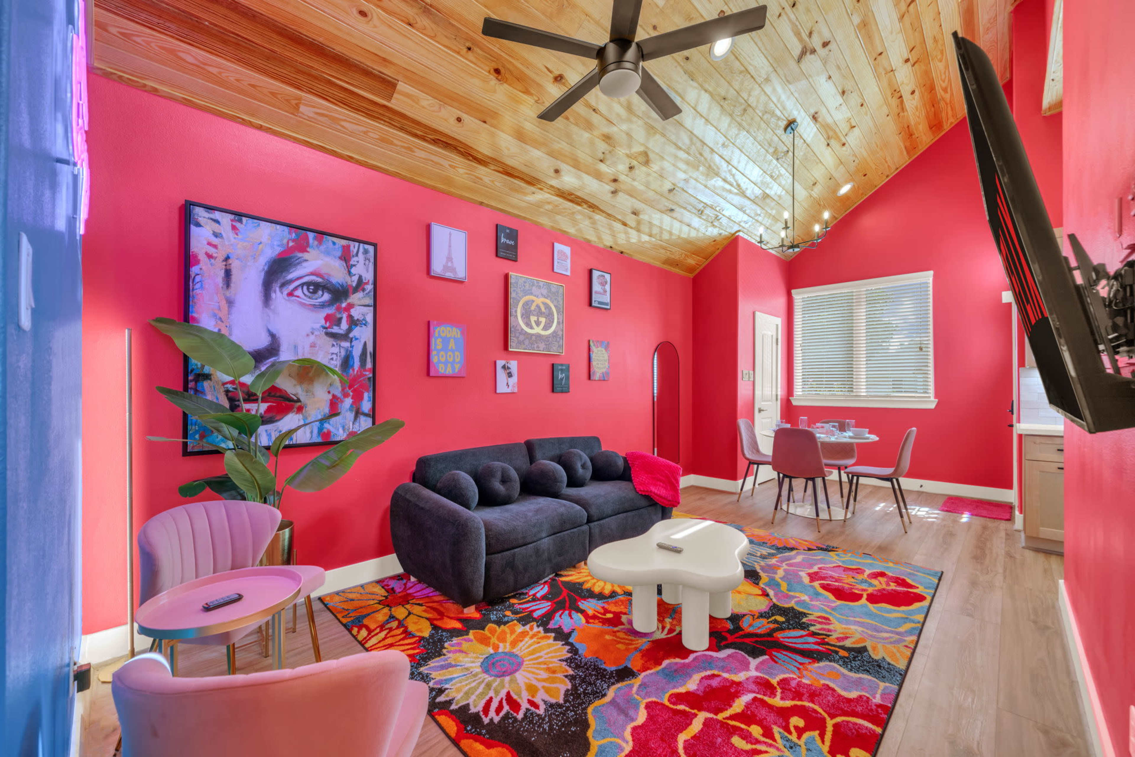 The room features a vibrant red wall with wooden ceiling beams, a dark sofa, colorful artwork, and a floral-patterned rug.