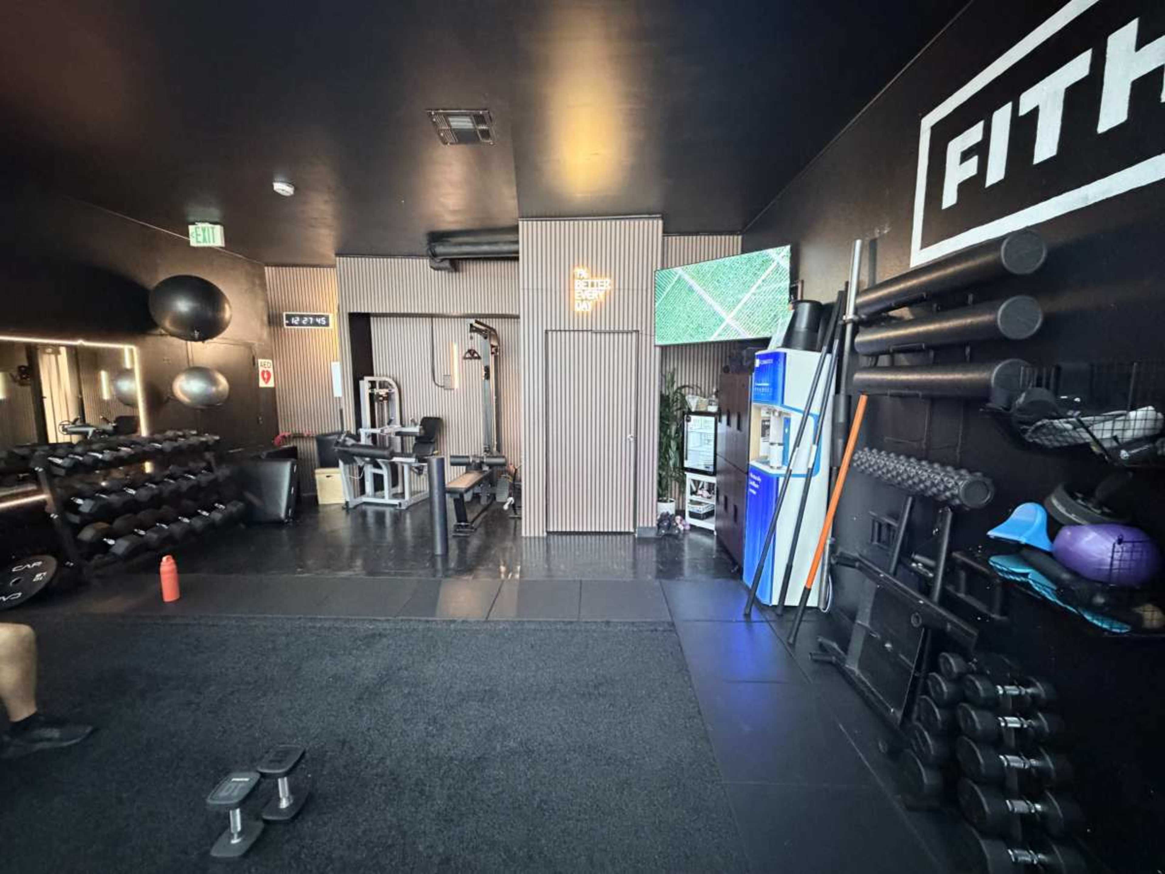 The image shows a modern gym interior with various workout equipment, including dumbbells and a weight bench, as well as a television displaying exercise content.