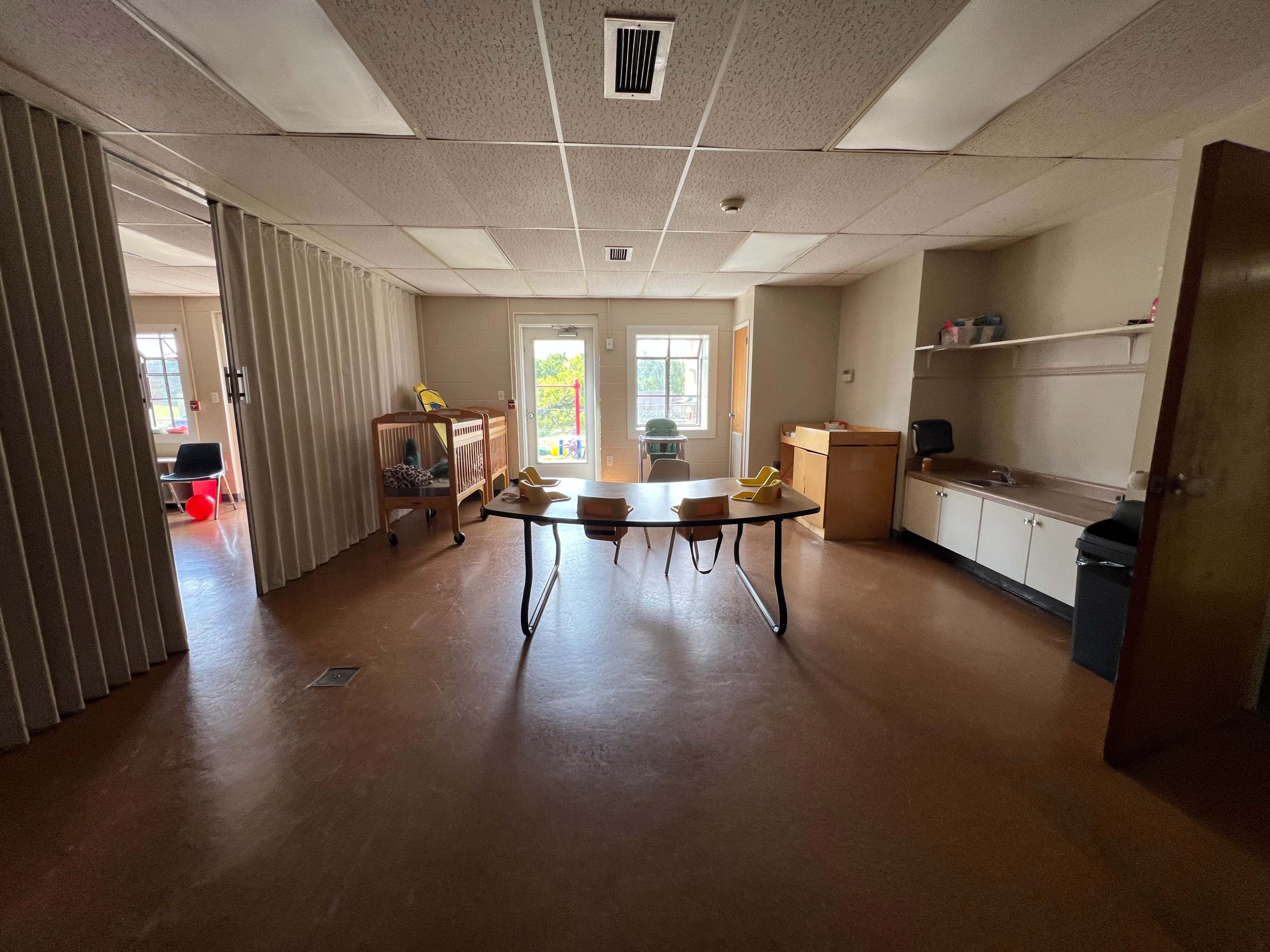 The image shows an empty room with a central table and chairs, surrounded by light-colored walls and a window that opens to an outdoor view.