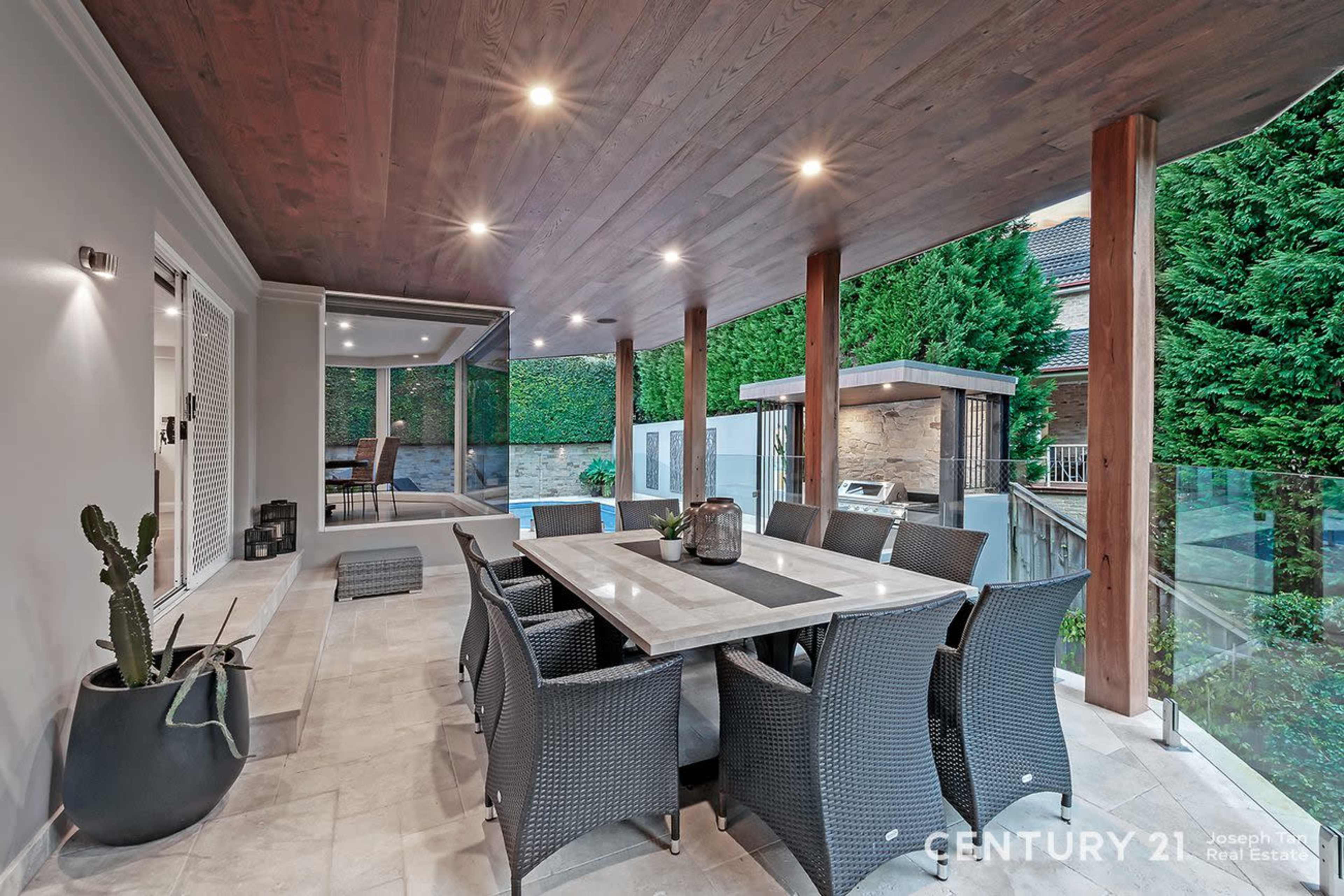 The image shows a modern patio with a dining table surrounded by wicker chairs, complemented by a stone wall and lush greenery in the background.