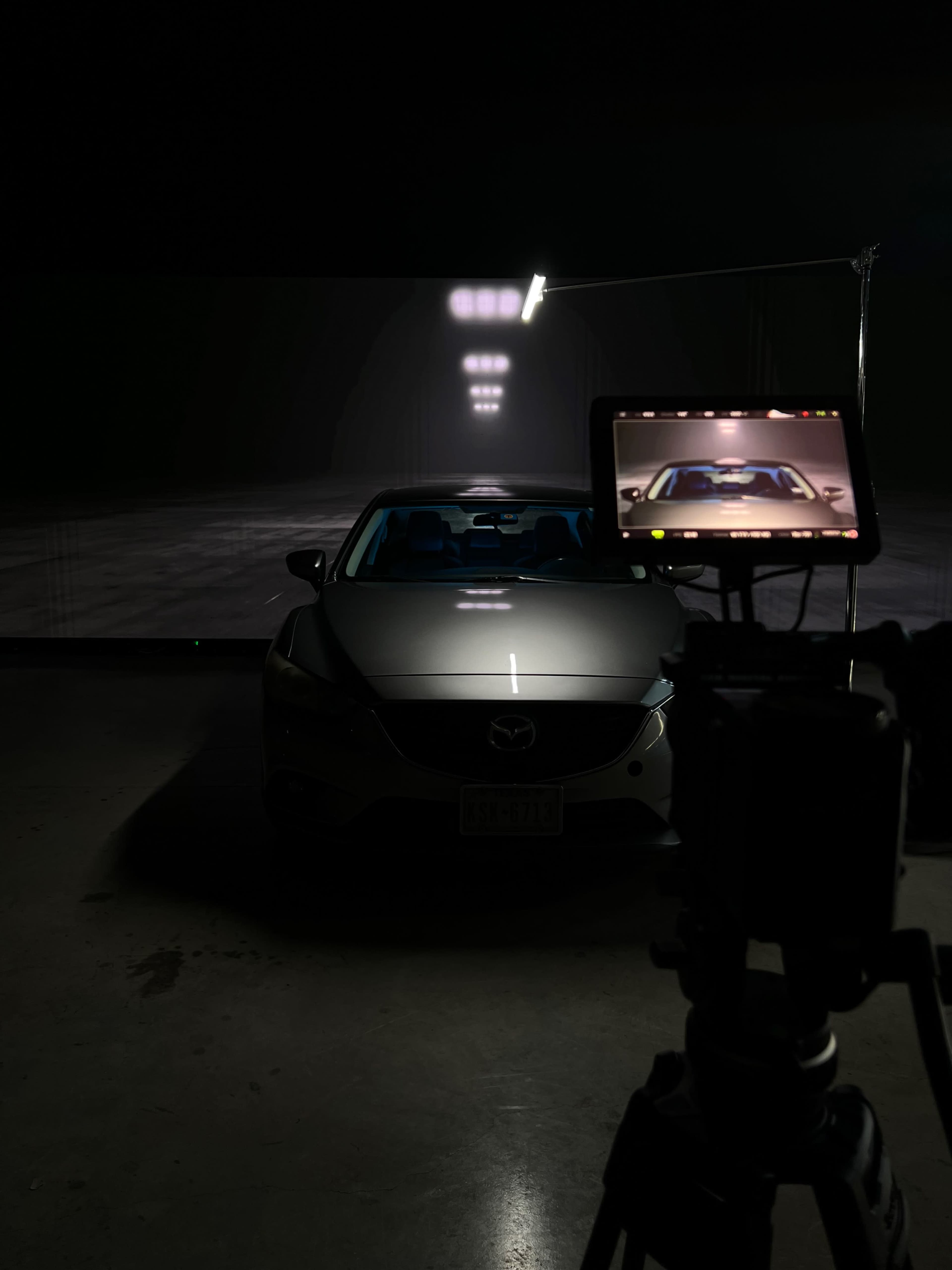 A silver Mazda car is positioned in a dimly lit studio with a camera setup in the foreground capturing the scene.
