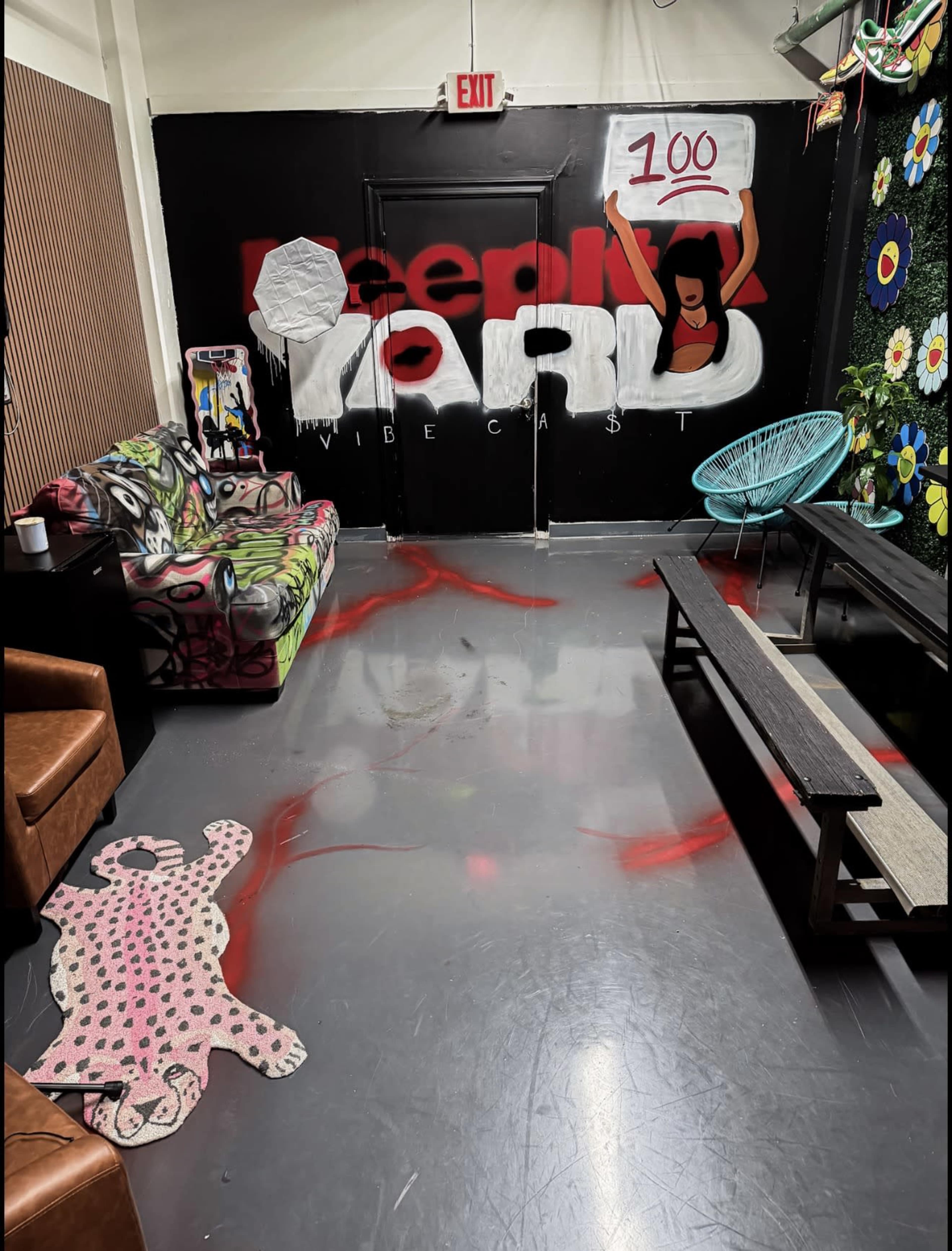 The image shows a colorful indoor space with graffiti on the wall, a graffiti-decorated couch, a patterned rug, and a wooden table against a vibrant backdrop of floral decorations.