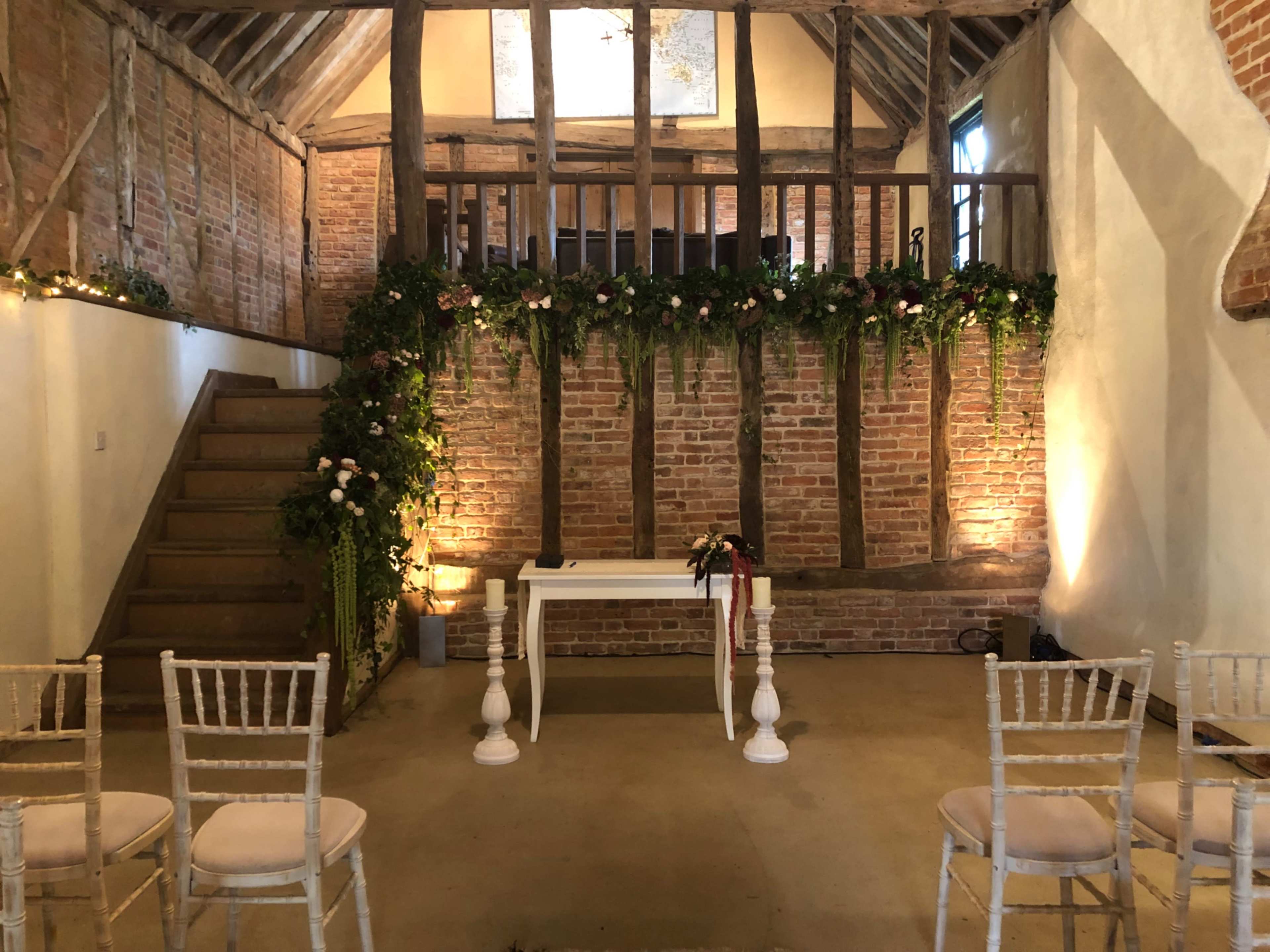 The image shows a rustic venue with exposed brick walls, a wooden staircase, and a decorated space for an event, featuring floral arrangements and chairs set up for guests.