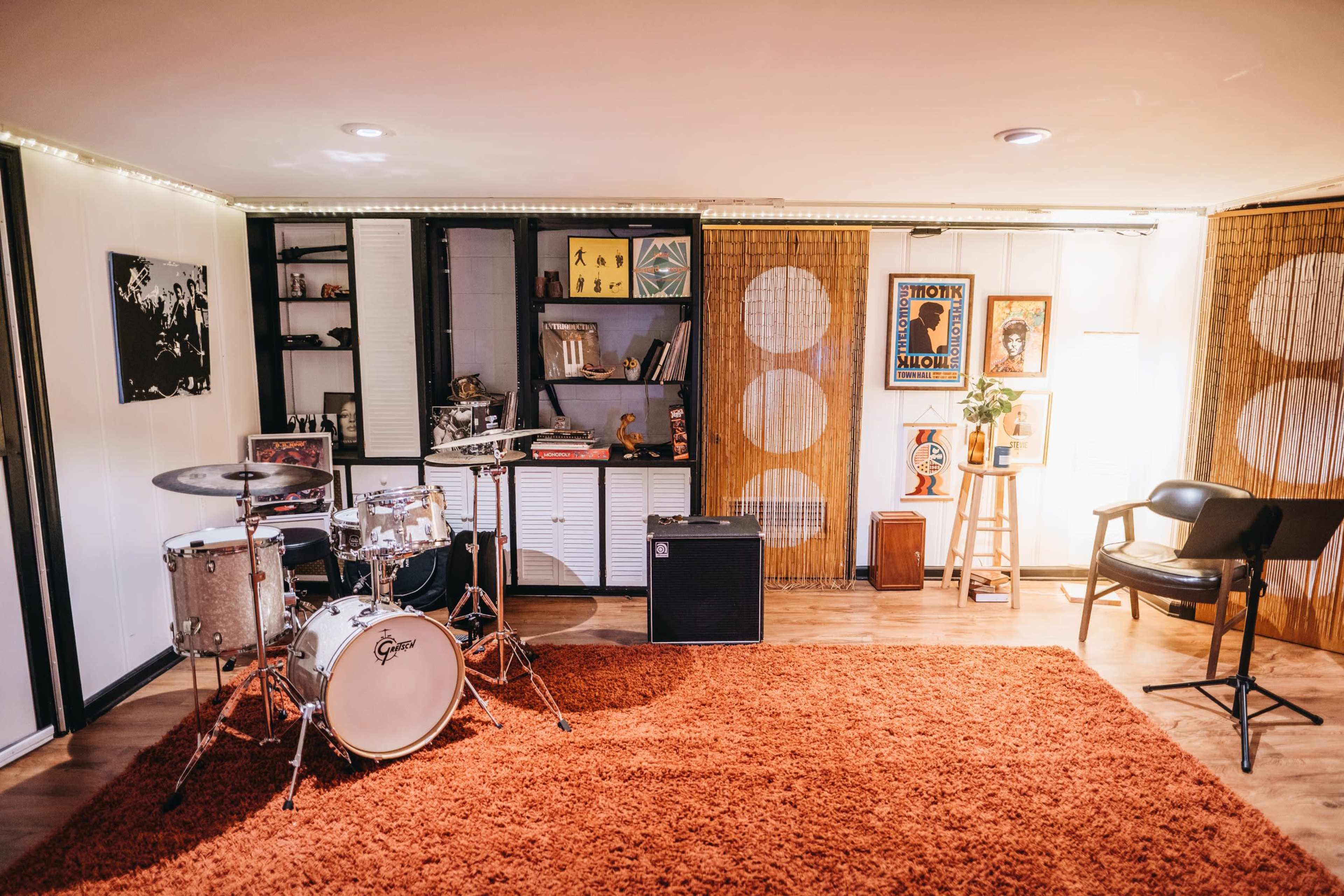 A music rehearsal space featuring a drum set, an amplifier, a bookshelf, and artwork displayed on the walls, all set on a large orange carpet.