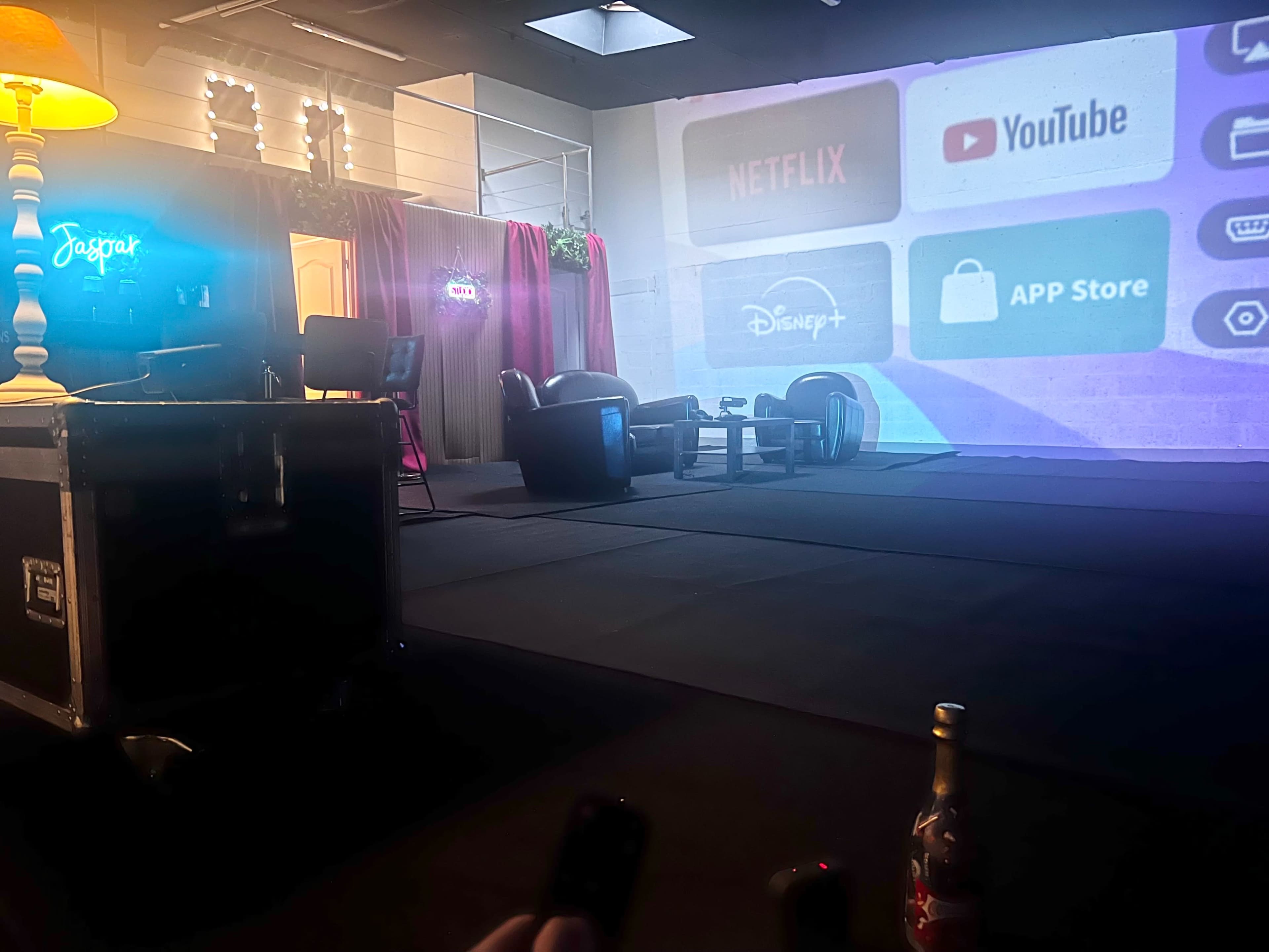 The image shows a dimly lit stage setup with a large screen displaying streaming app logos, two armchairs facing each other, and a decorative lamp.