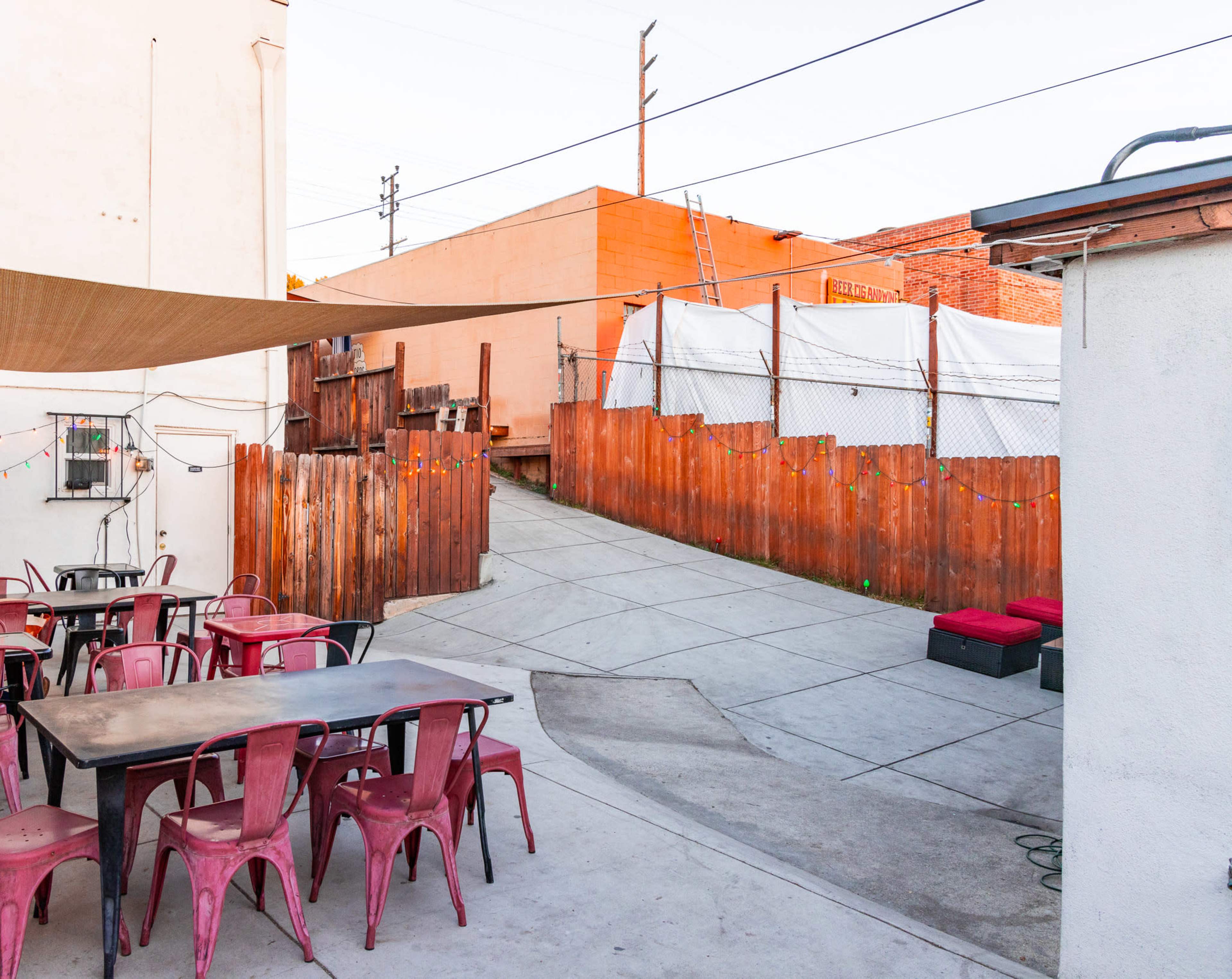 A narrow alleyway is bordered by wooden fences and leads toward a building, with metal tables and chairs arranged in the foreground.