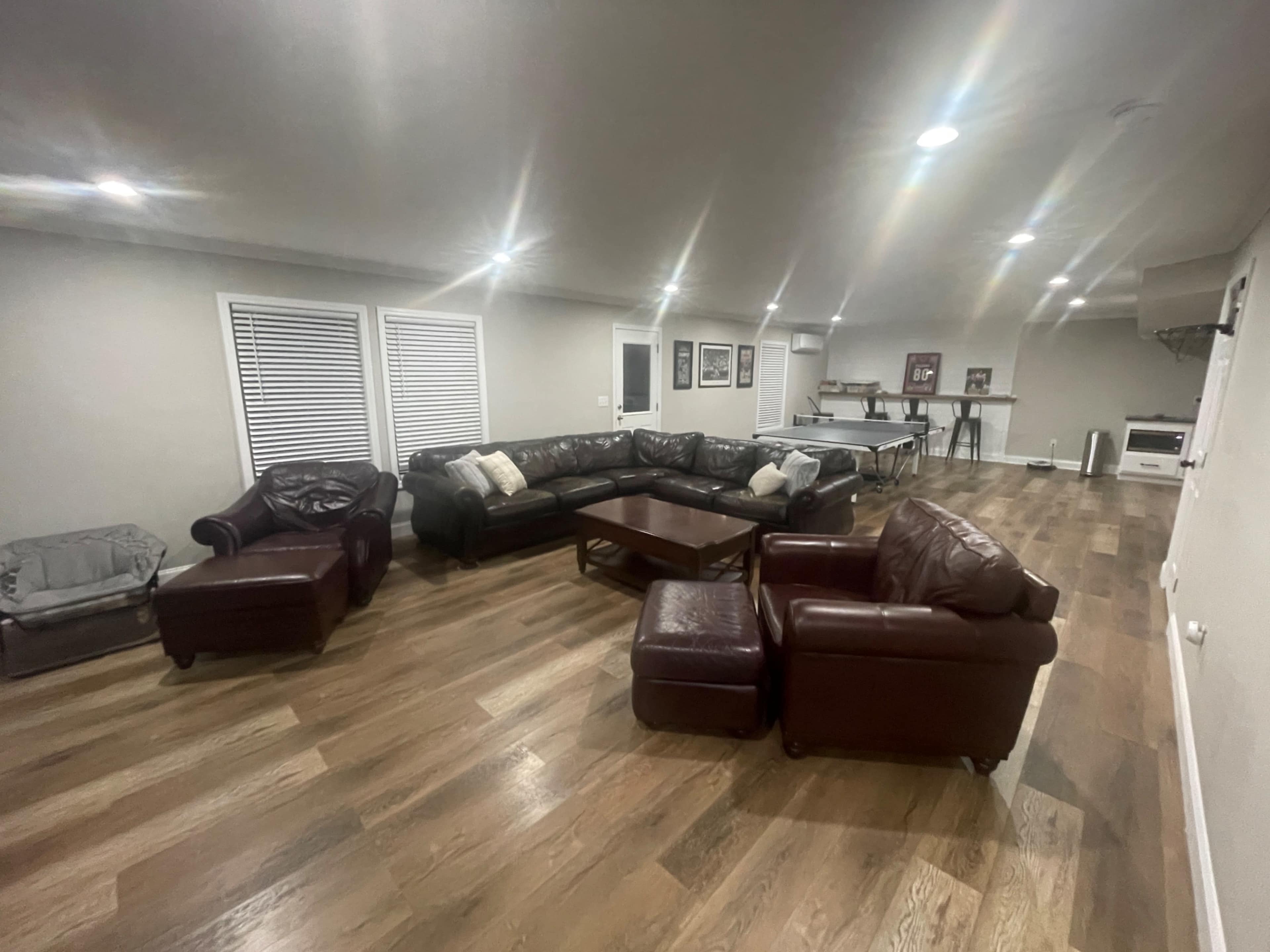 The image shows a spacious living room featuring a large sectional couch, two armchairs, a coffee table, and hardwood flooring, with windows providing natural light.