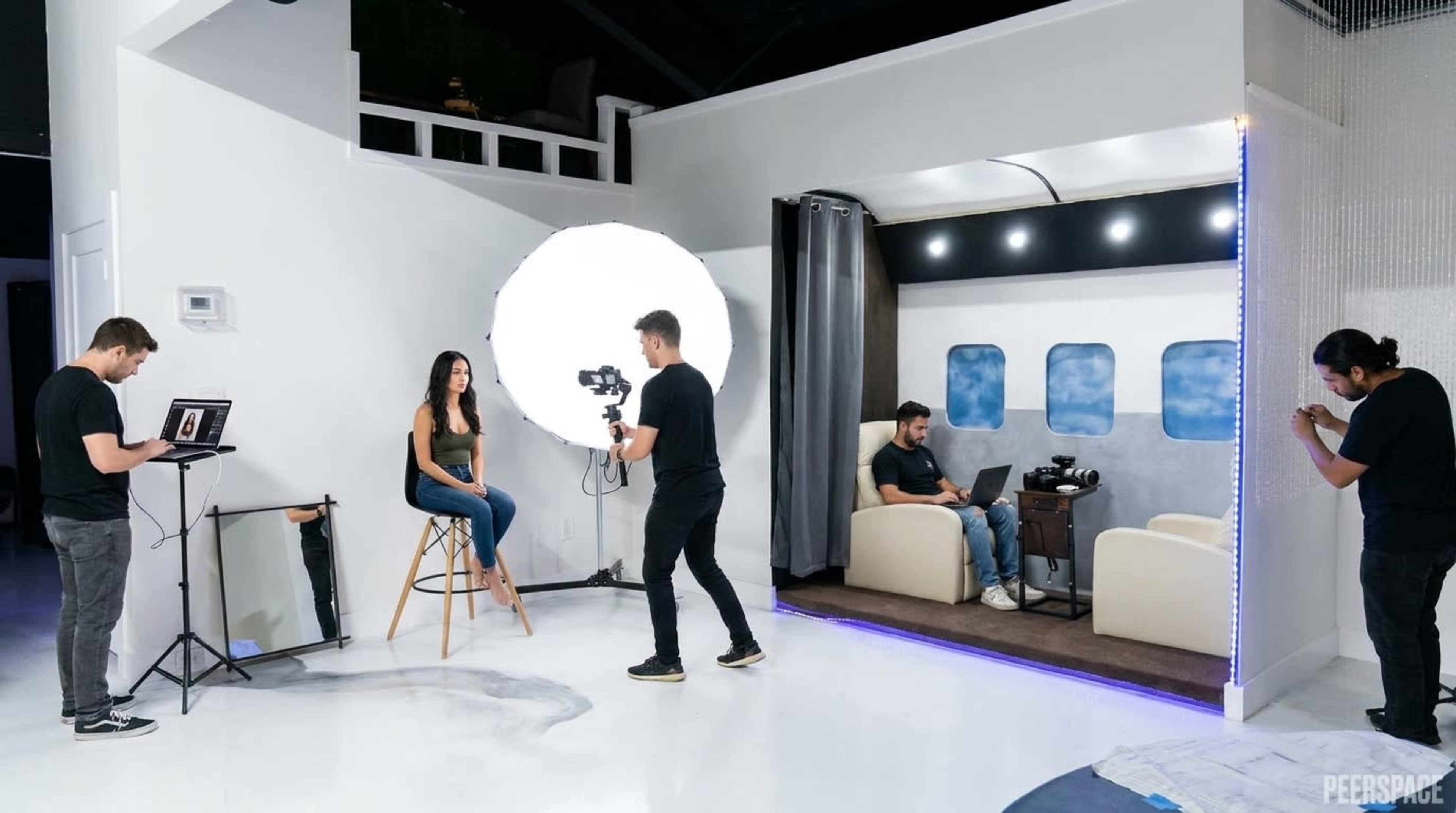 A photoshoot is taking place in a studio featuring a model on a stool, a large lighting setup, and several photographers and crew members engaged in their work around a set designed like an aircraft interior.