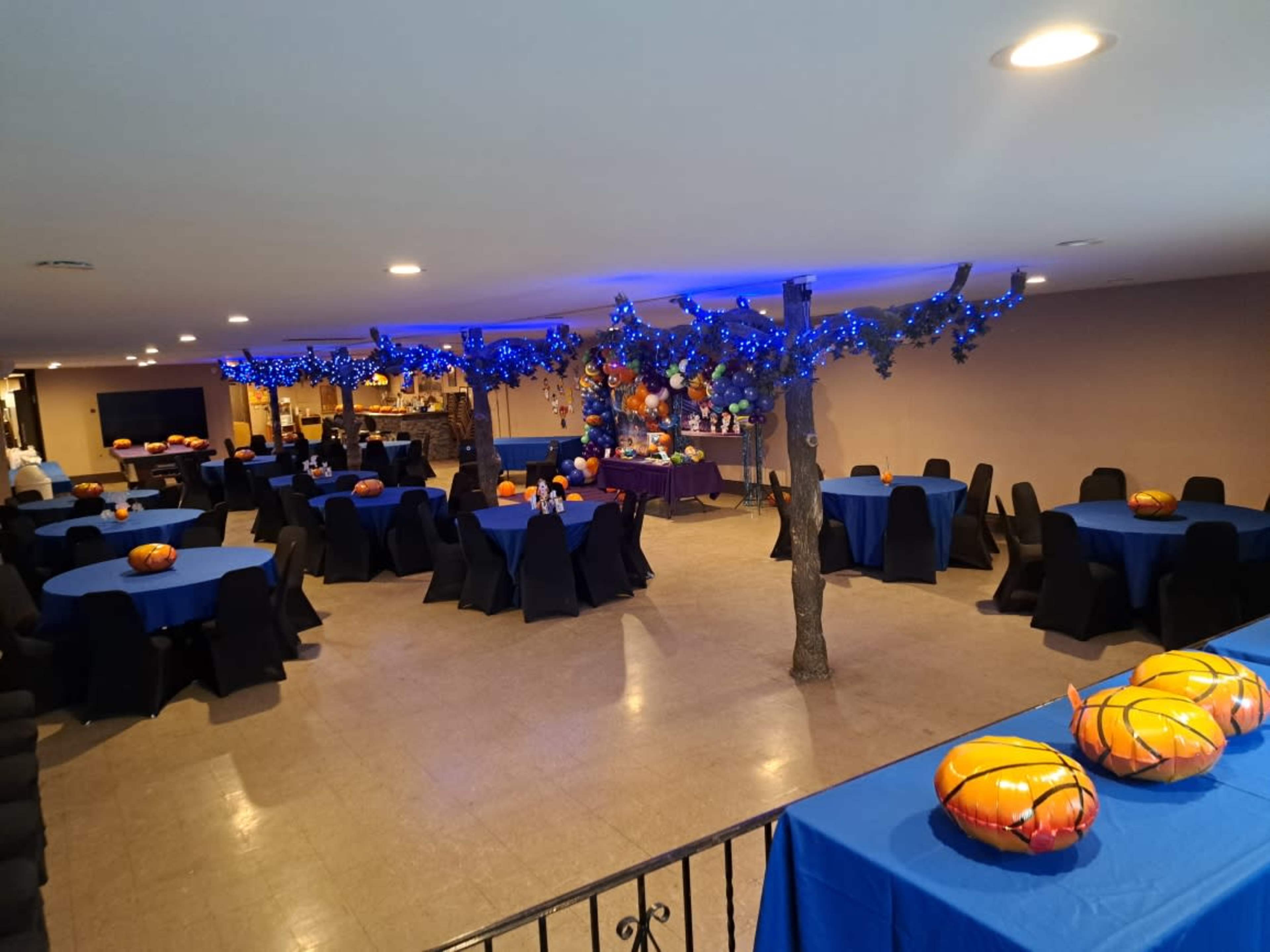 A decorated event space with blue tablecloth-covered tables, black chairs, and orange pumpkin centerpieces, all under blue string lights.