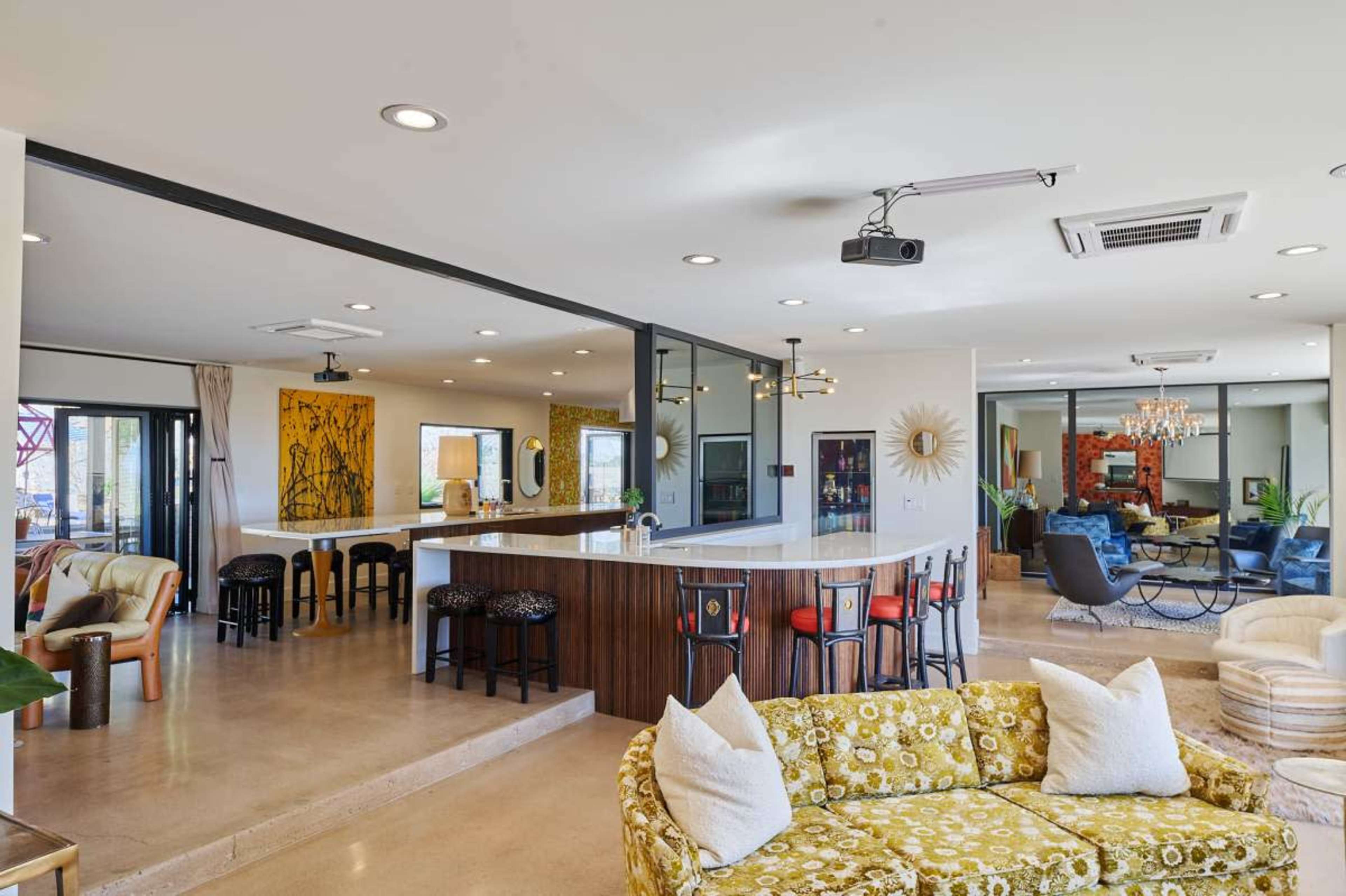 The image shows a modern living space with a kitchen bar, a seating area featuring a patterned sofa, and large windows allowing natural light.